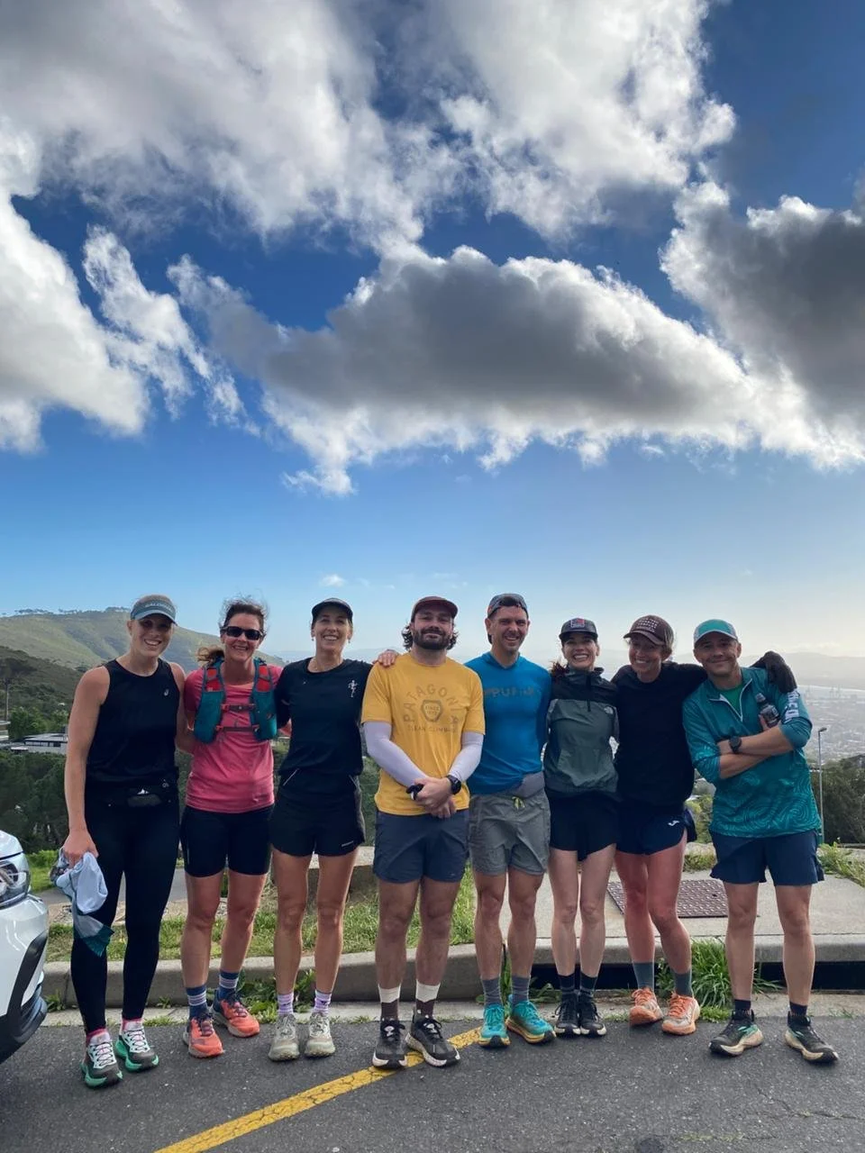 Group of nine people dressed in athletic clothing, standing arm in arm outdoors on a mountain trail, with cloudy sky and scenic landscape in the background.