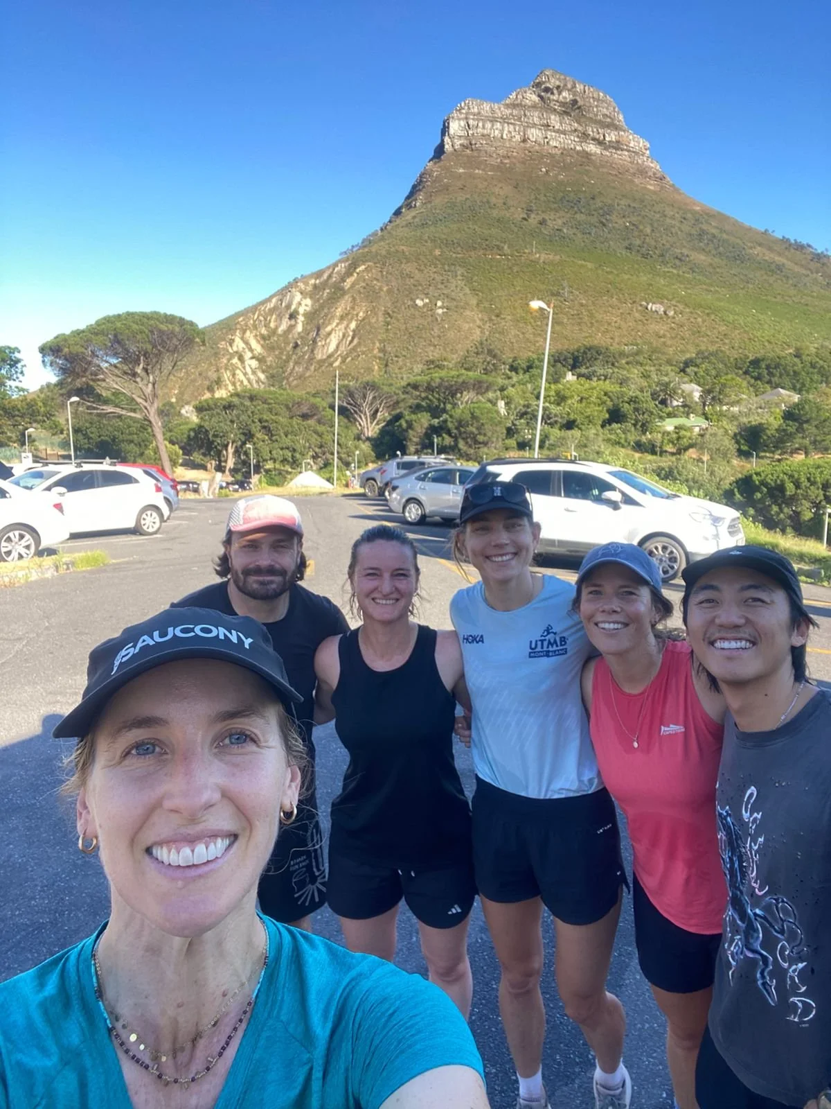 Group of six runners taking a selfie with a mountain in the background, parked cars, and trees in a parking lot on a sunny day.
