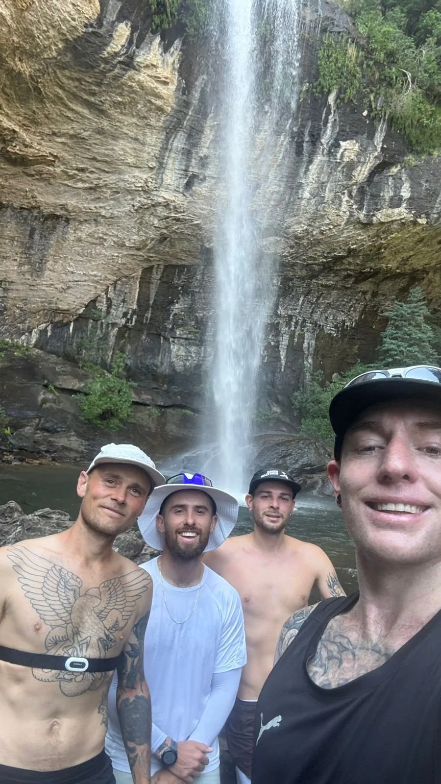 Four men posing in front of a waterfall with water cascading down a cliff in a lush forest setting.