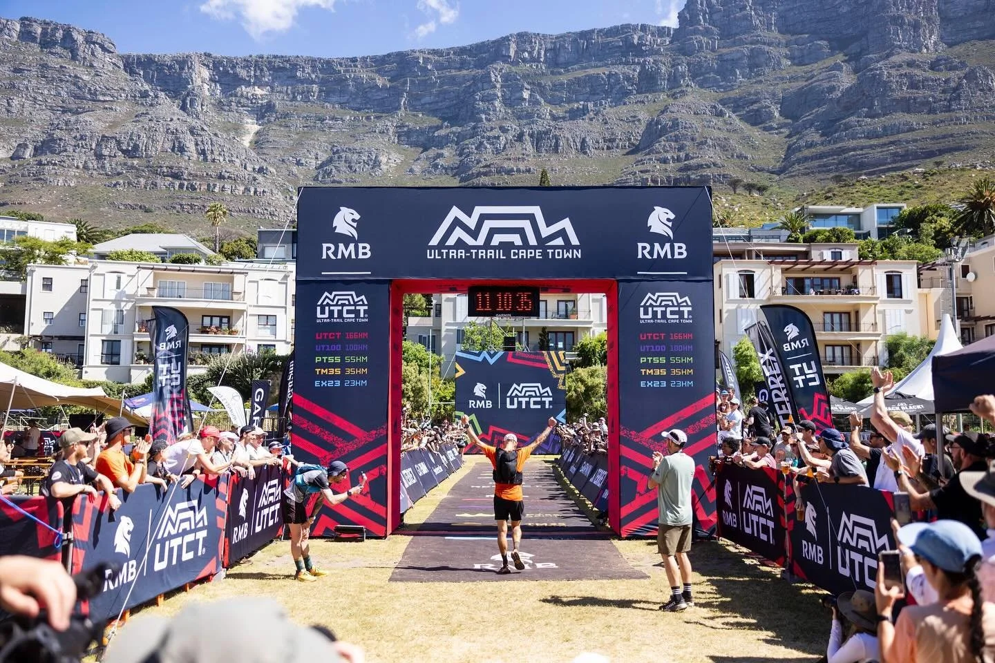 Runner crossing the finish line at the Ultra-Trail Cape Town race, surrounded by spectators on a sunny day with mountainous landscape in the background.