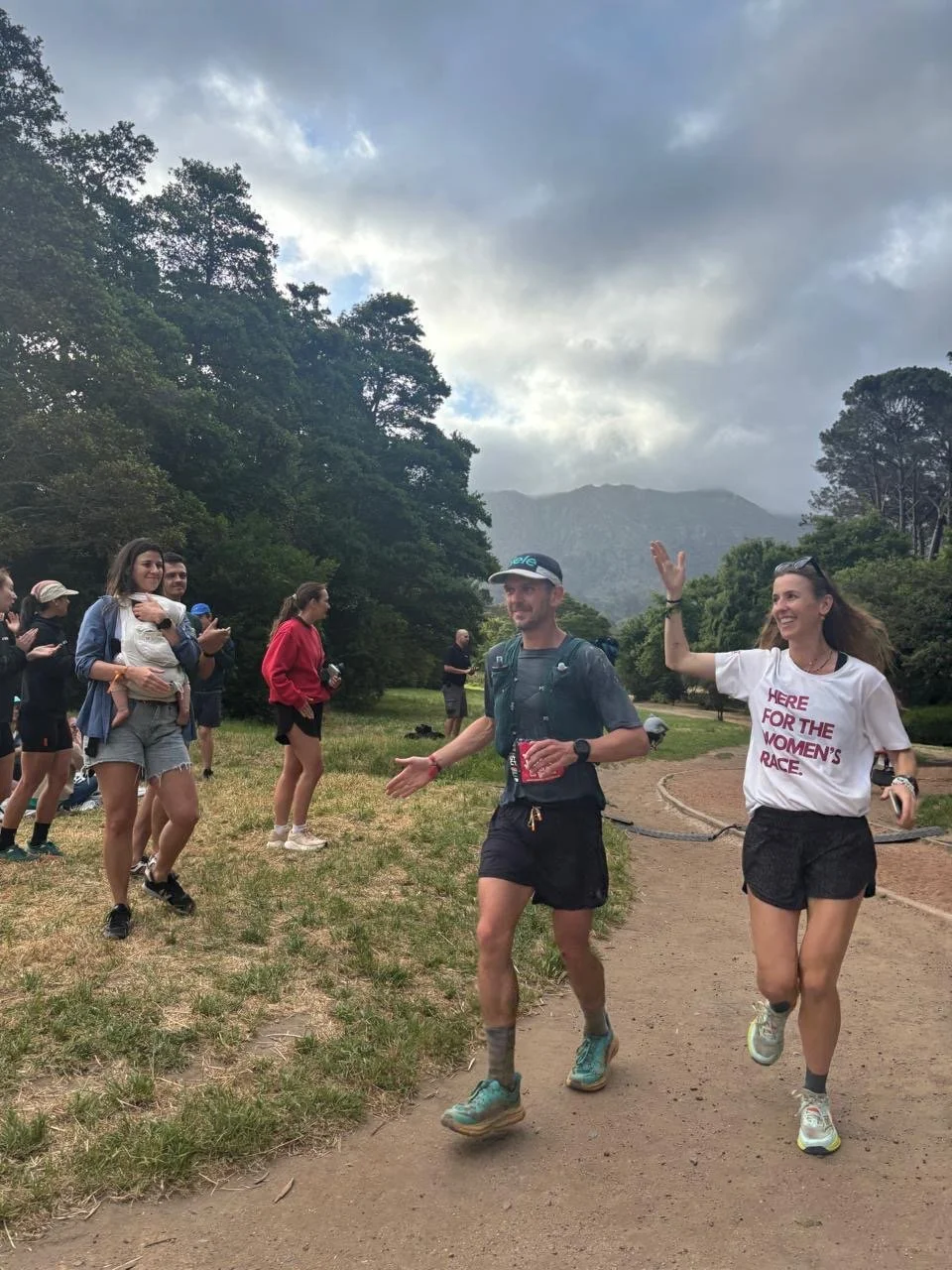 People running and cheering on a dirt trail in a scenic outdoor area with trees and mountains, cloudy sky.
