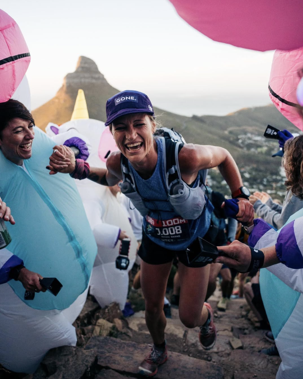 Female runner participating in a mountain race, smiling and being cheered on by supporters dressed in unicorn costumes, with a mountain in the background at sunrise or sunset.