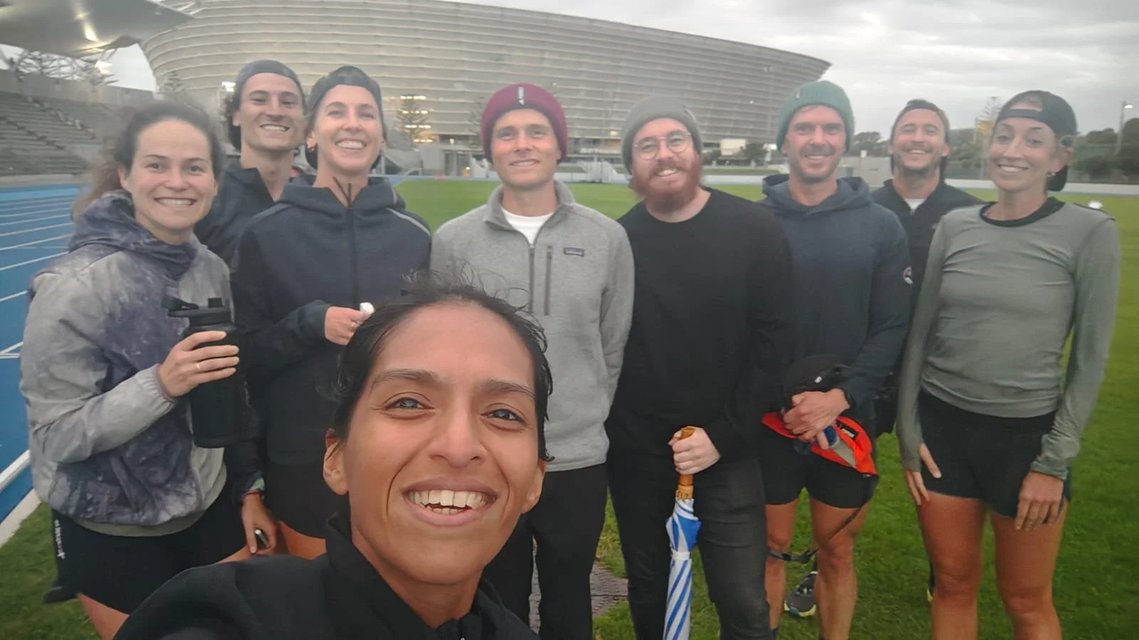 Group of nine runners smiling and posing for a photo at an outdoor track and stadium.