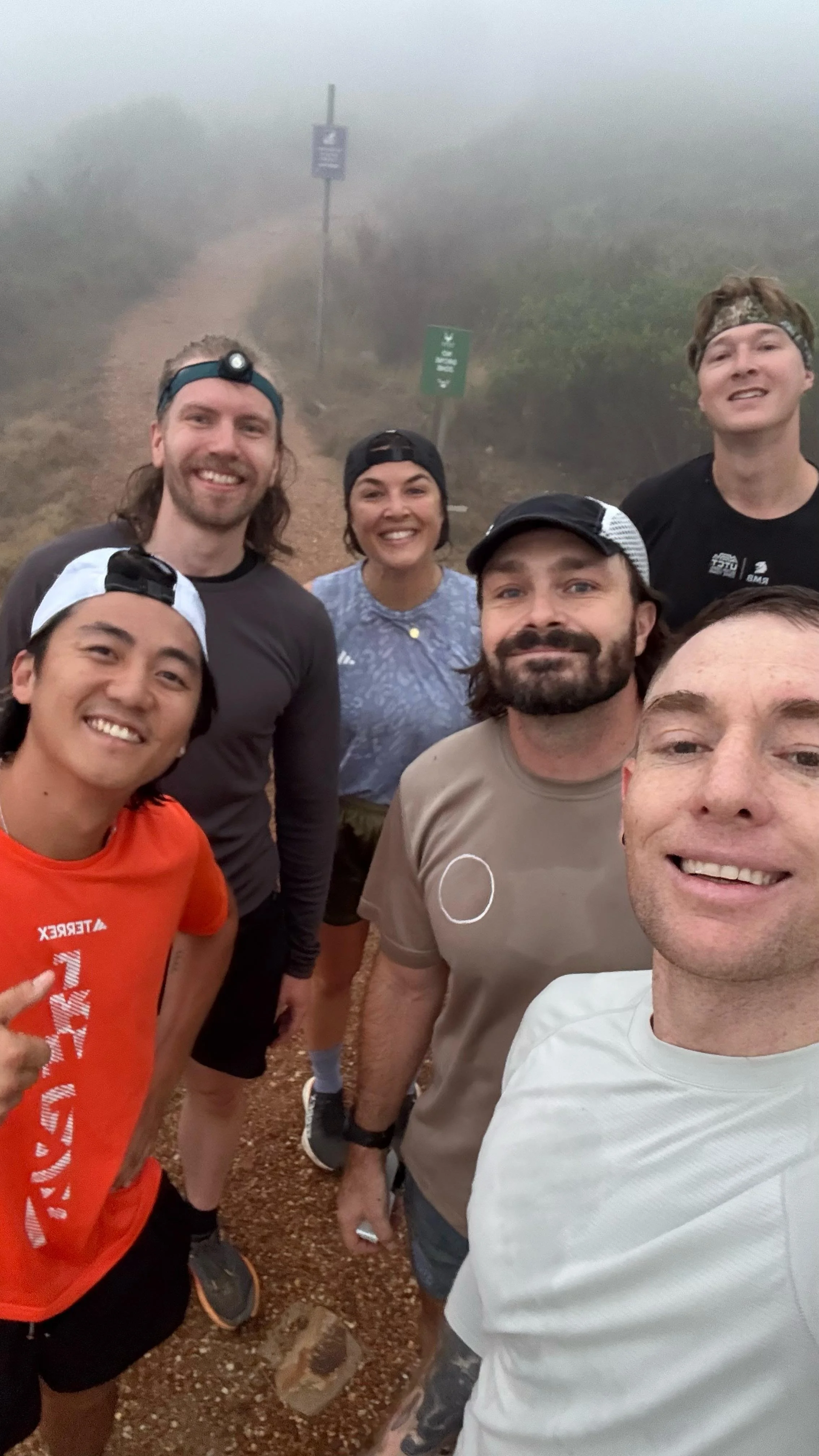 Group of six smiling people taking a selfie on a foggy trail during a hike.