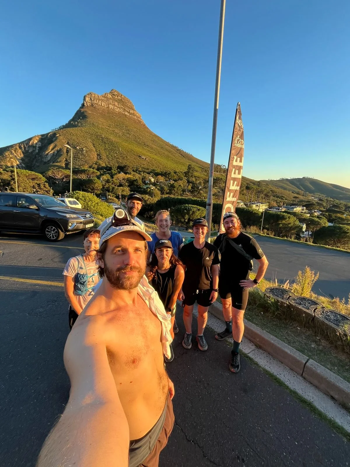 Group of seven runners in athletic gear taking a selfie outdoors in front of a mountain at sunset.