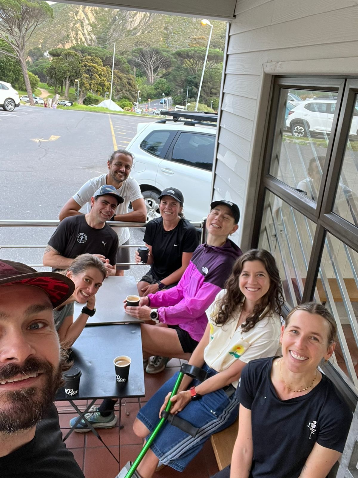 A group of smiling people sitting at an outdoor cafe table, holding cups of coffee, with mountains and trees in the background.