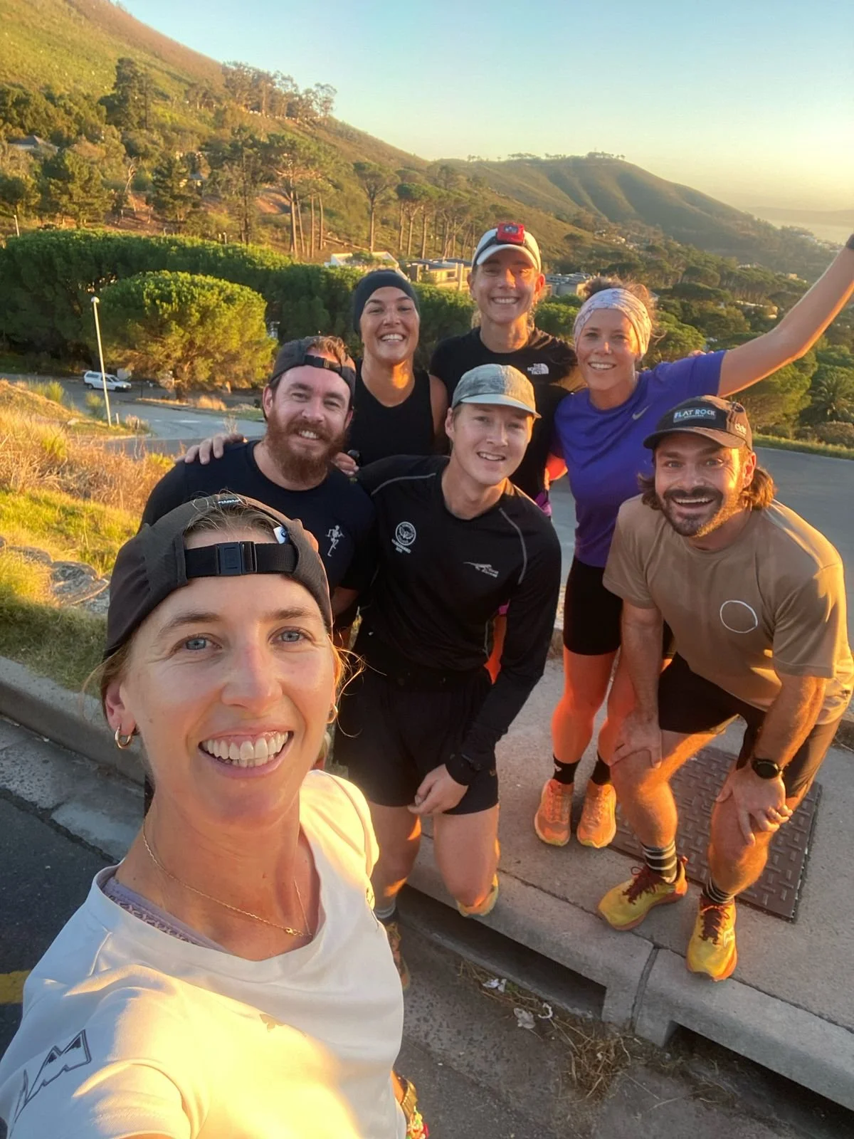 Group of seven runners taking a selfie outdoors during sunset, with green hills and trees in the background.