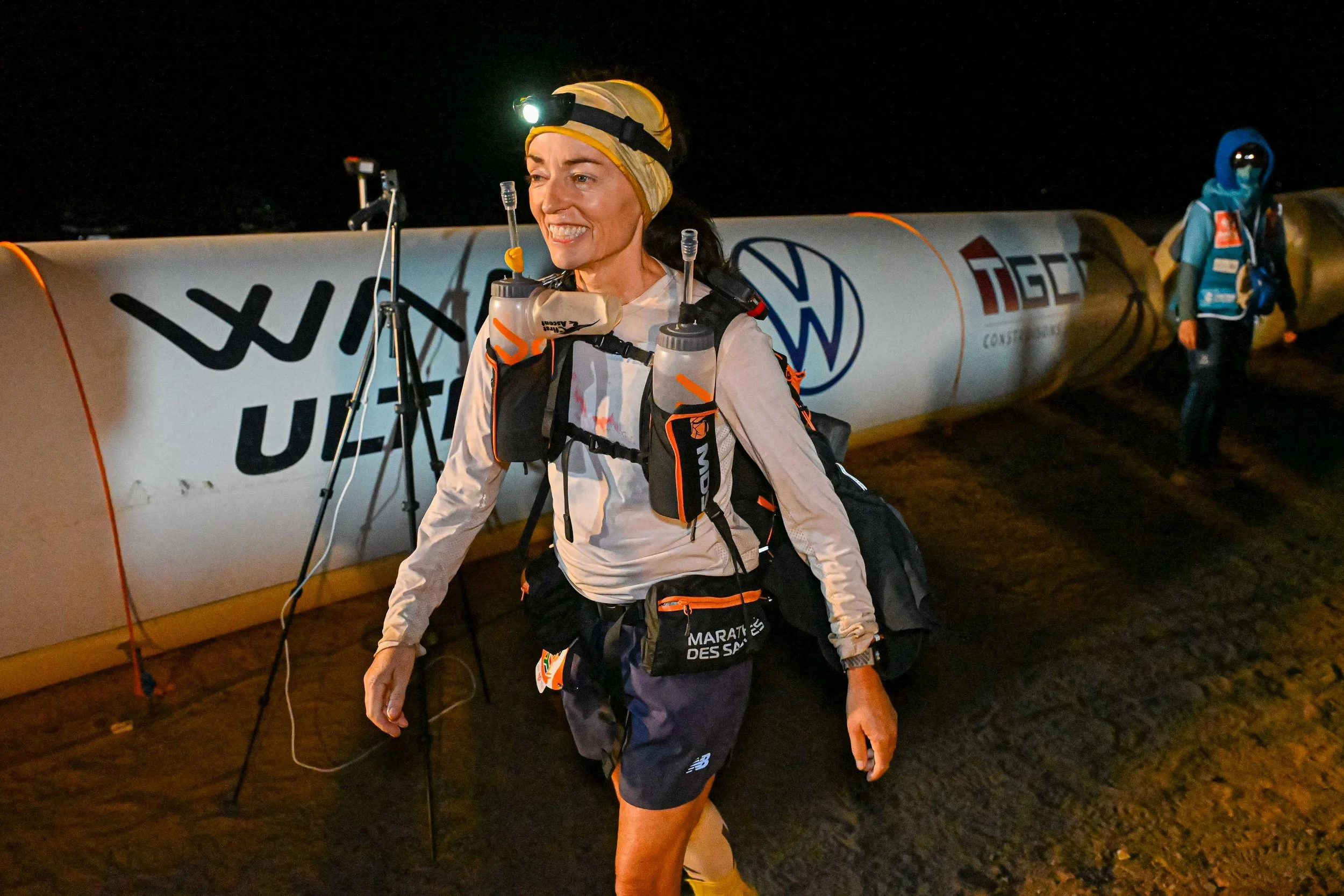 A woman in athletic gear with a headlamp, carrying two water bottles, smiling, standing in front of a large inflatable arch at night, with another person in the background.