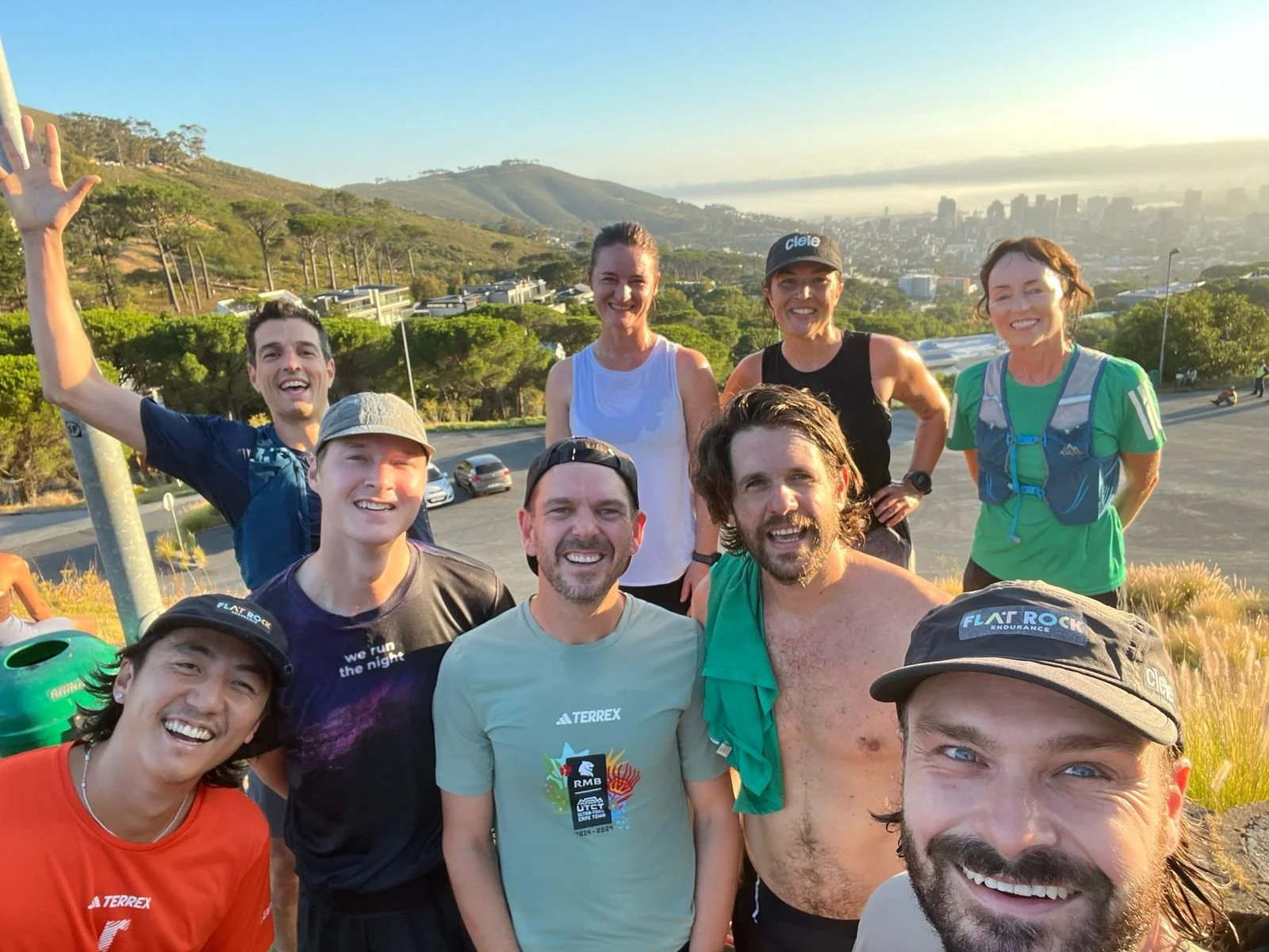 A group of nine people smiling outdoors during the daytime, with hills and a cityscape in the background.