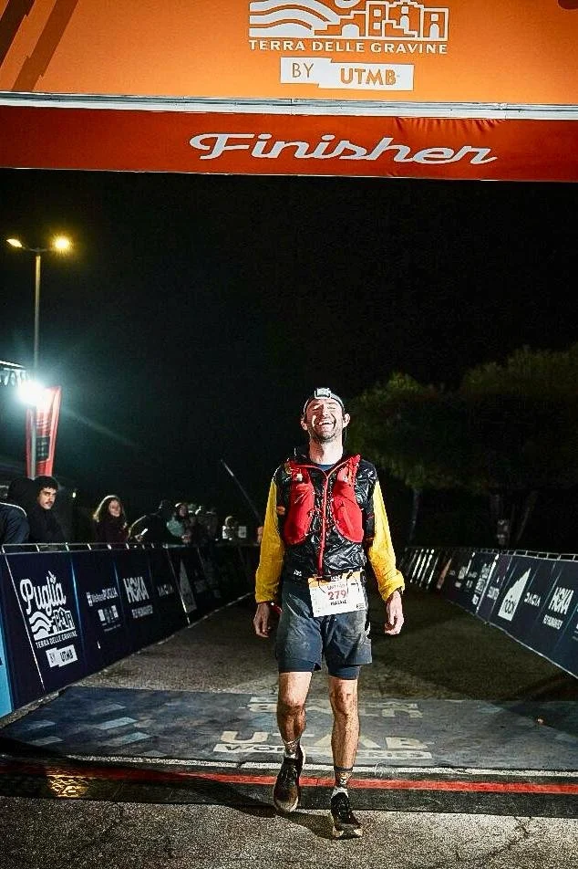 A man crossing the finish line at a nighttime race event, smiling and wearing athletic gear and a bib number 279