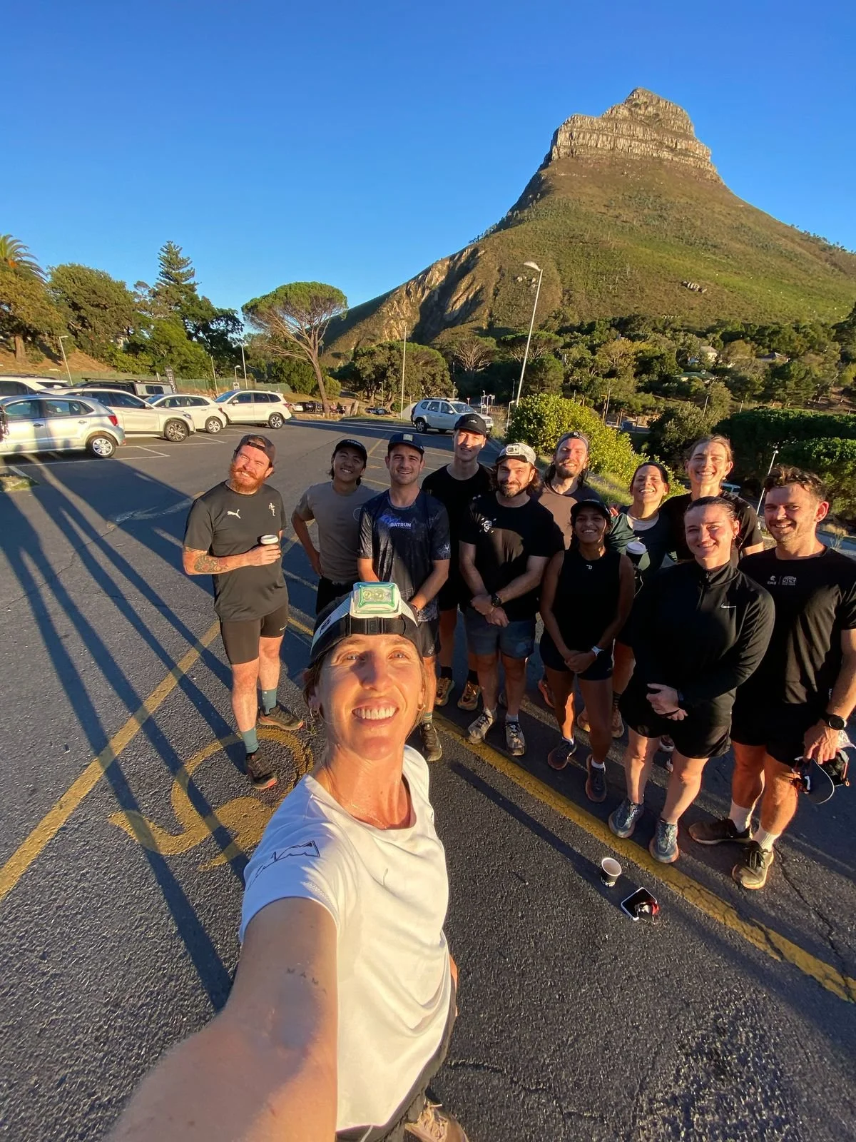 A group of people in athletic gear posing for a selfie in a parking lot with a mountain in the background during sunset.