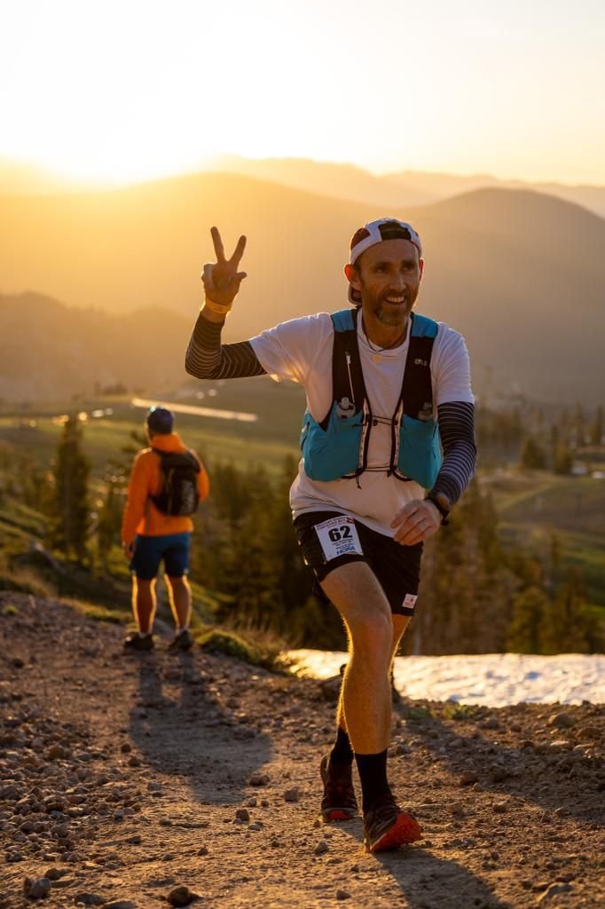 Man running on trail during sunset, making a peace sign with his hand, wearing a headband and a hydration backpack.