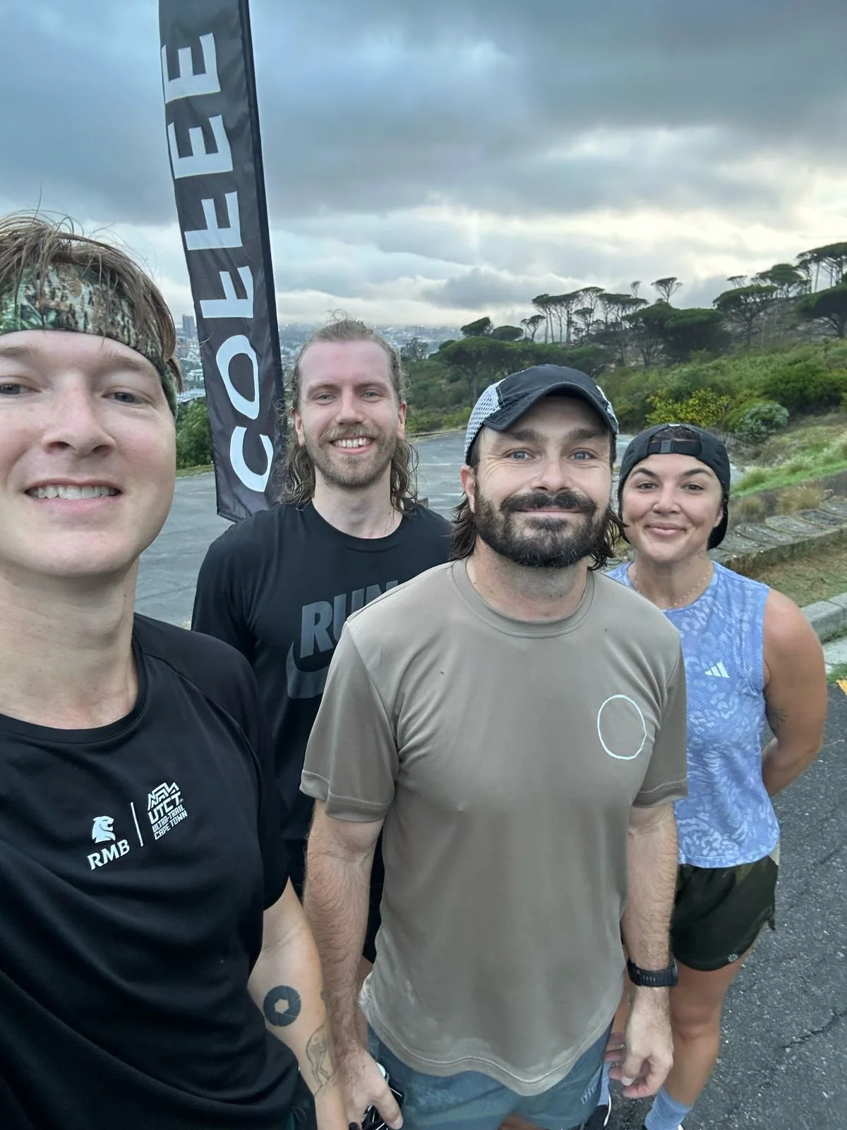 Four people taking a selfie outdoors with a cloudy sky and trees in the background. They are dressed in athletic clothing and appear to be participating in a running event, with a flag that says 'CRREE' next to them.