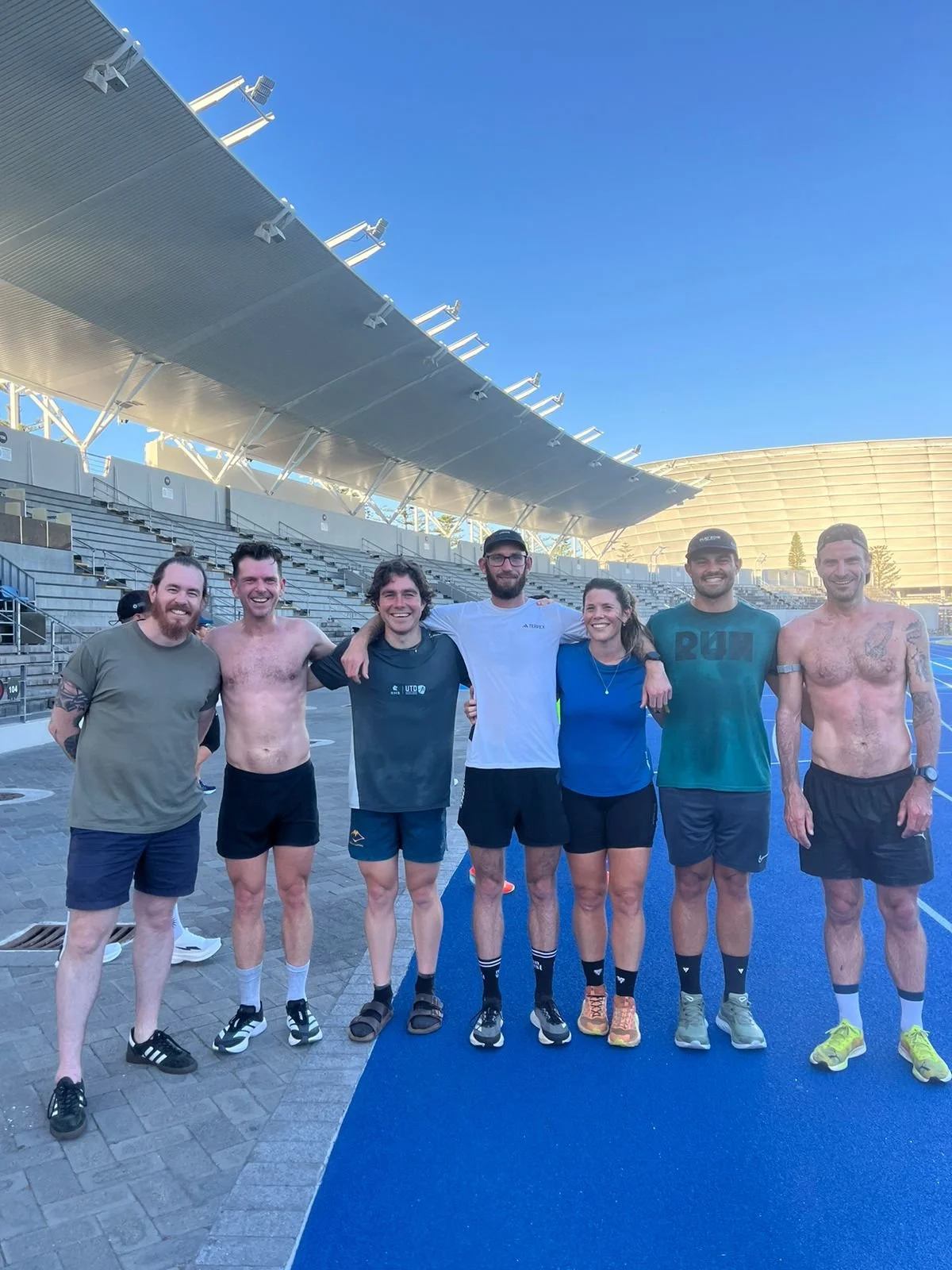 A group of seven people standing together on a blue running track, smiling with arms around each other, in an outdoor stadium with stands and a large canopy overhead, during daytime.