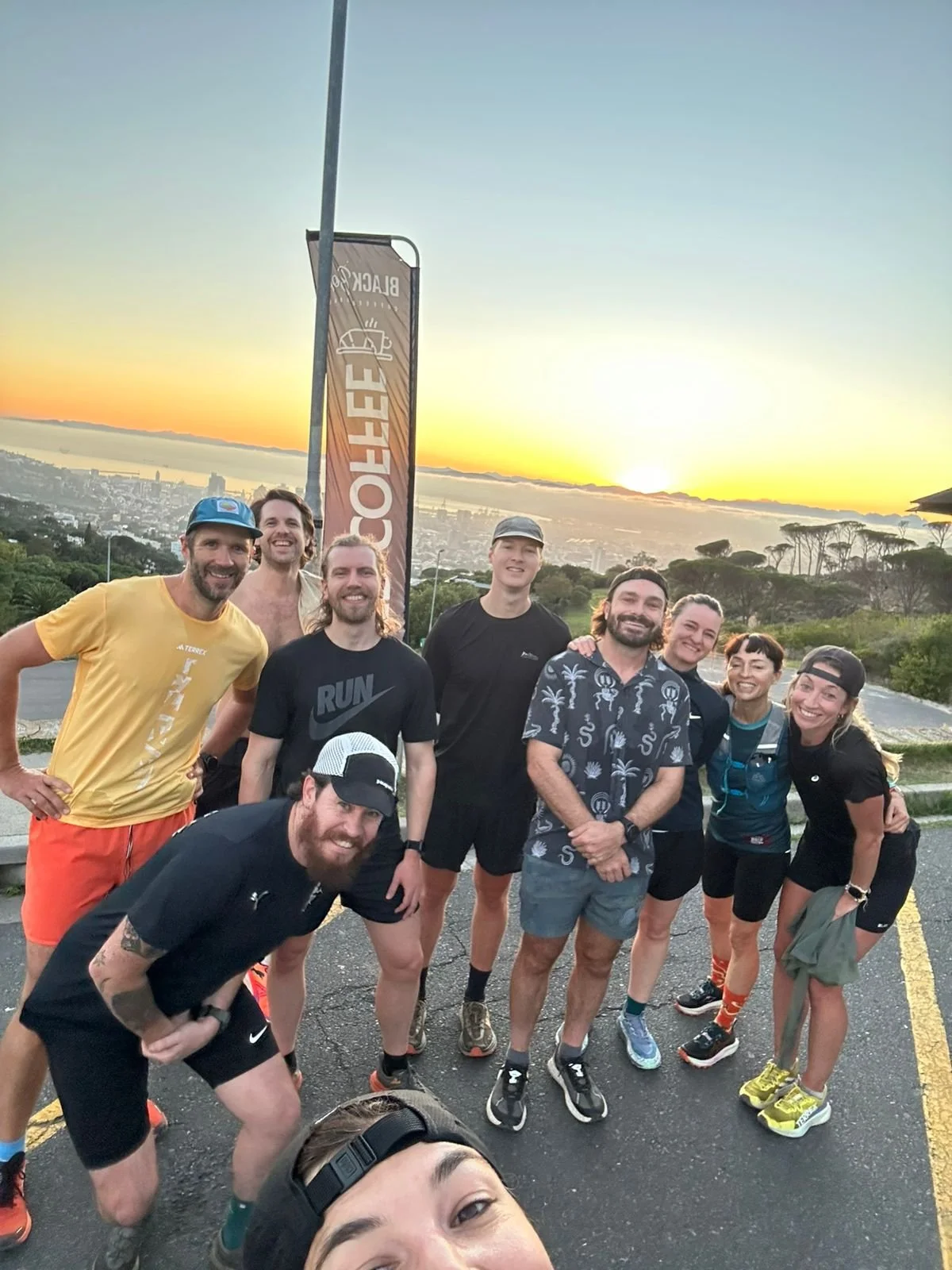 A group of nine people, dressed in athletic clothing, standing together outdoors at sunrise with a city skyline and fog in the background, posing for a photo after a run.