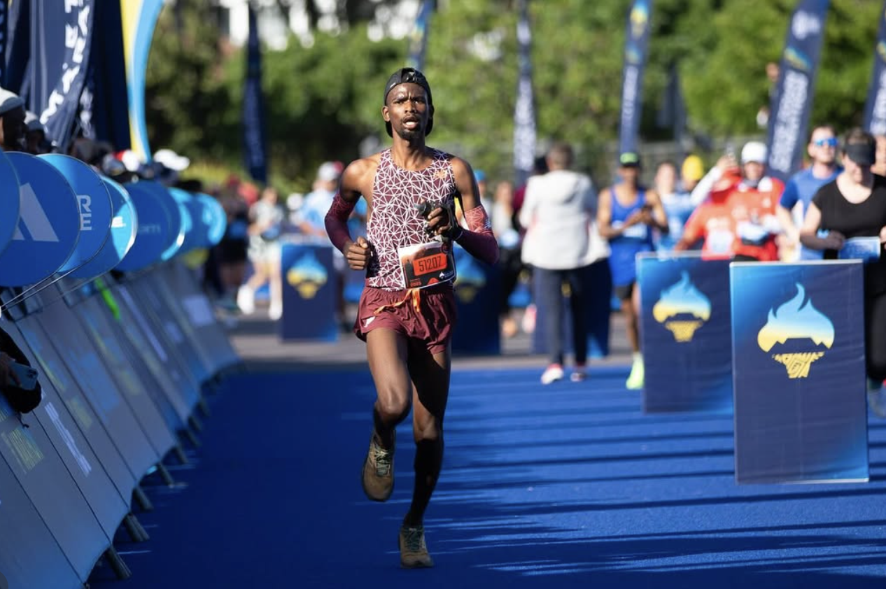A male marathon runner in a maroon and purple athletic outfit running on a blue track, with spectators and race officials in the background.