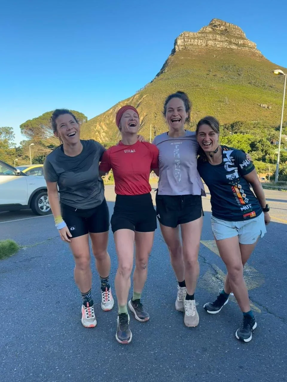 Four women in athletic clothing smiling and laughing outdoors with a mountain in the background.
