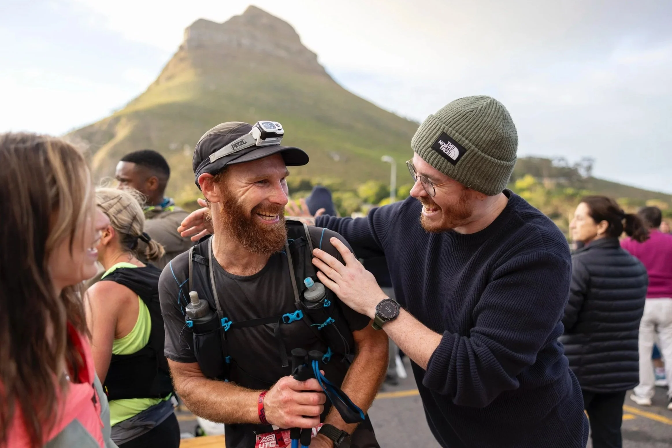 Two men sharing a joyful moment at an outdoor event, with a mountain in the background. One man has a beard, wearing a black shirt, a backpack, and a headlamp. The other man is wearing glasses, a green beanie, and a navy sweater, and is smiling as he touches the first man's shoulder.