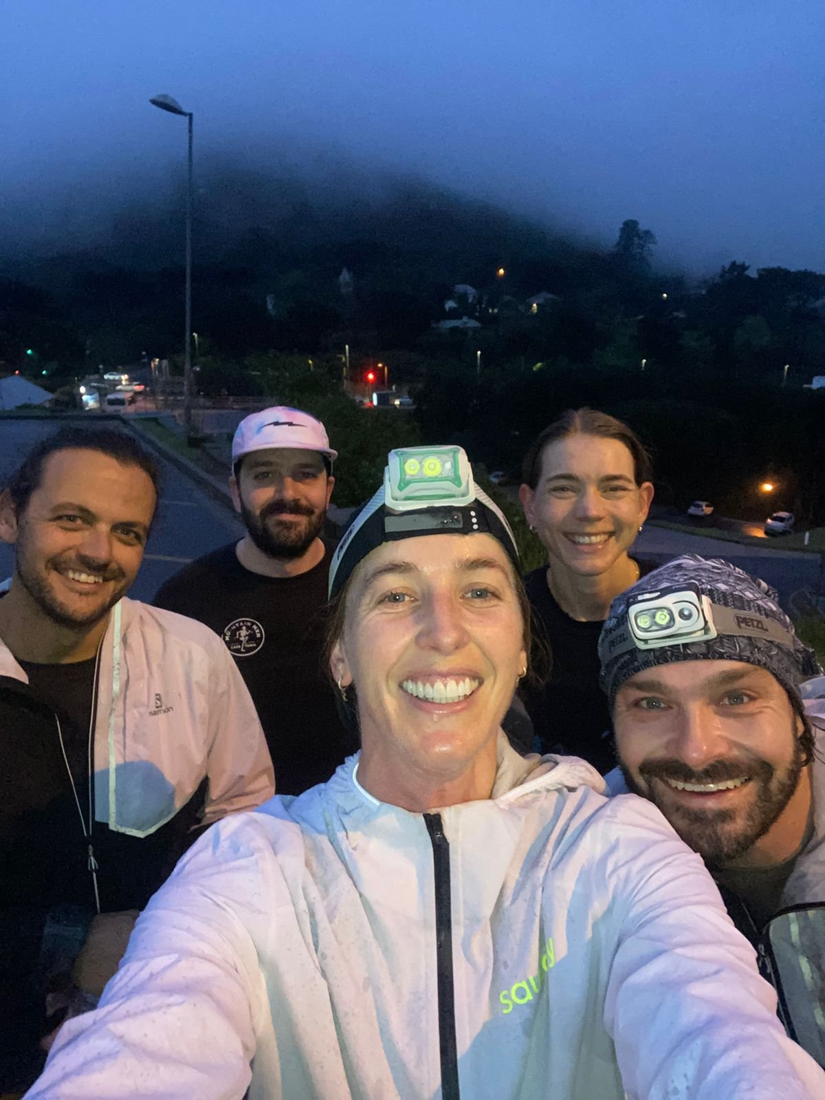 Group of five smiling friends taking a selfie outdoors in the evening with a dark sky and streetlights in the background