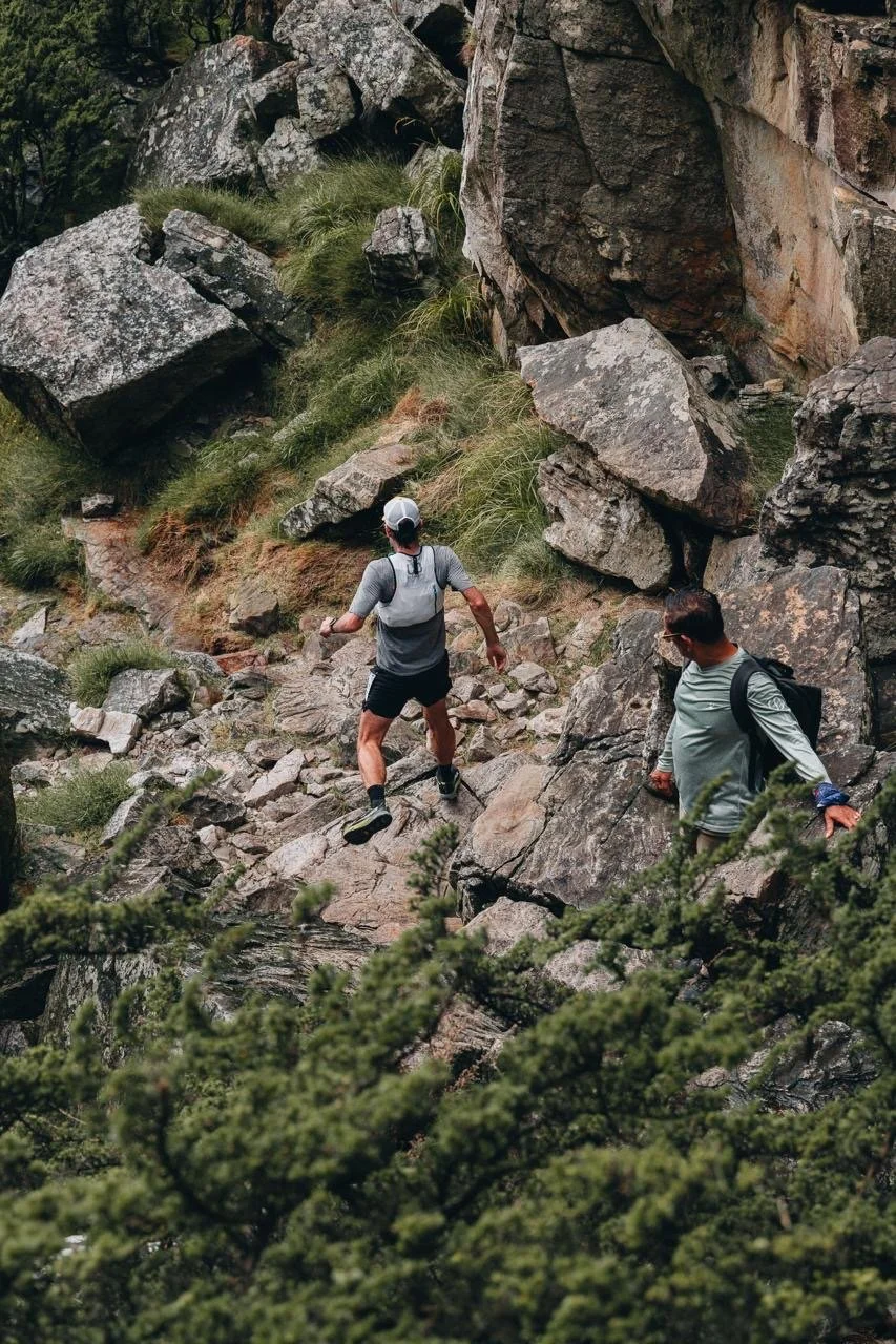 Two hikers walking on a rocky trail surrounded by large rocks and green vegetation.