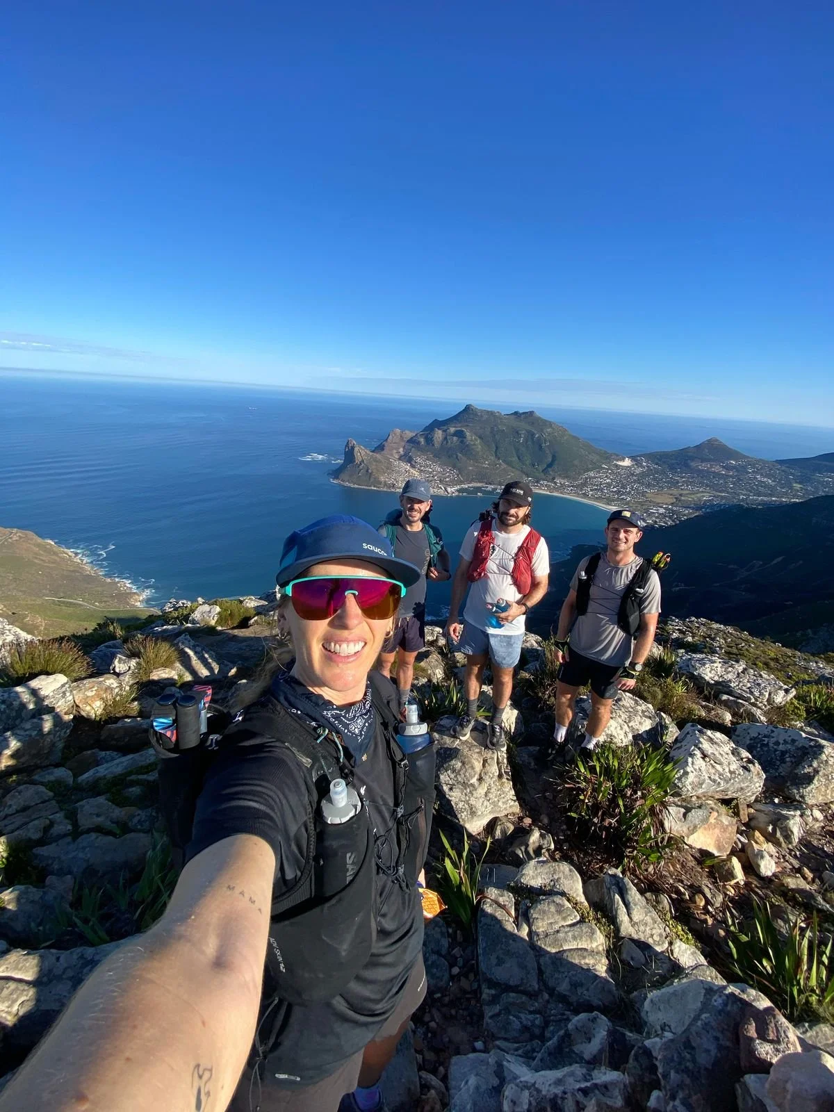 A group of five hikers taking a selfie at the top of a mountain with a view of the ocean and a mountainous island in the background on a sunny day.
