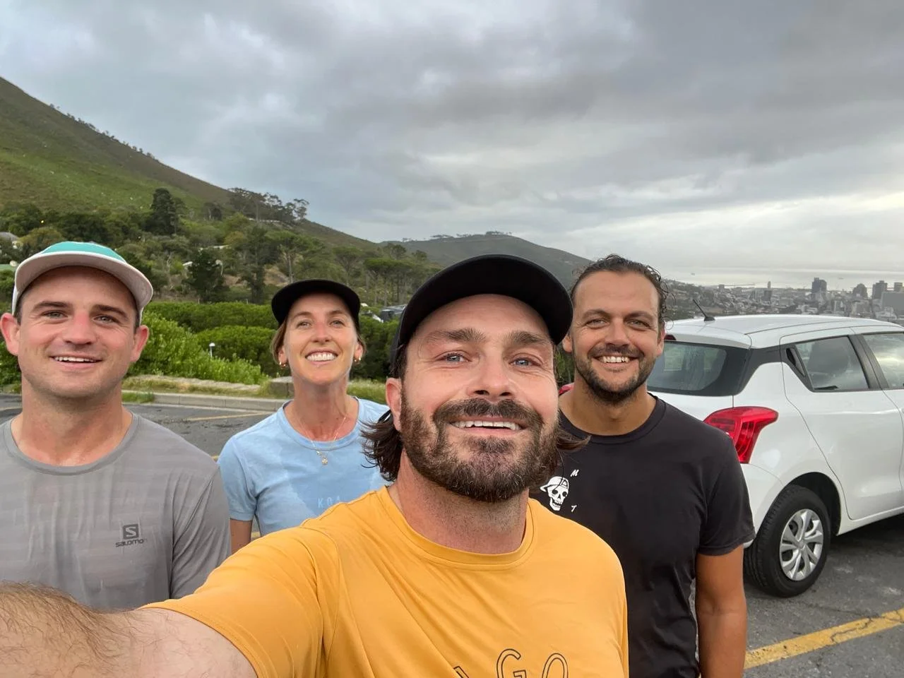 Four friends taking a selfie outdoors near a city and hillside, with a white SUV in the background, under cloudy skies.