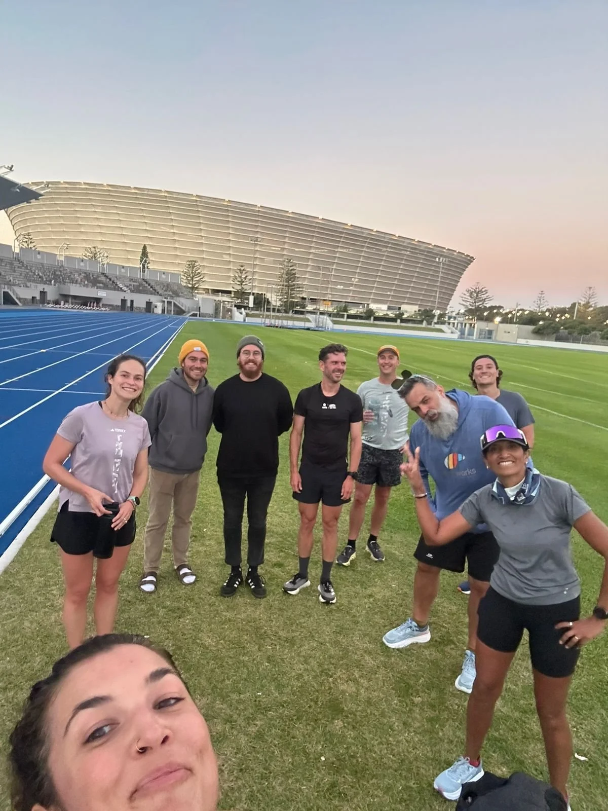 Group of people standing on a track field at an outdoor stadium at sunset, smiling and posing for a photo.