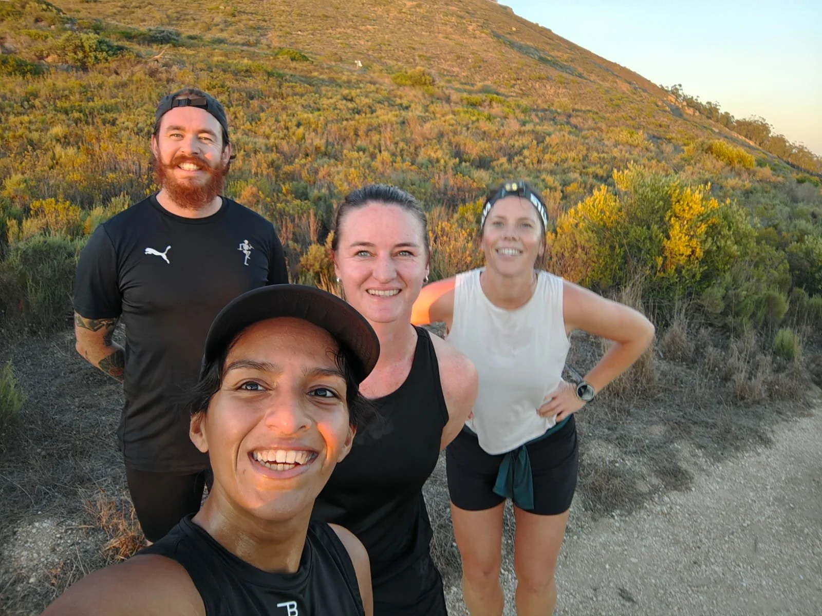 Four people taking a selfie outdoors on a trail with a hillside covered in bushes and trees in the background, during what appears to be sunset or sunrise.