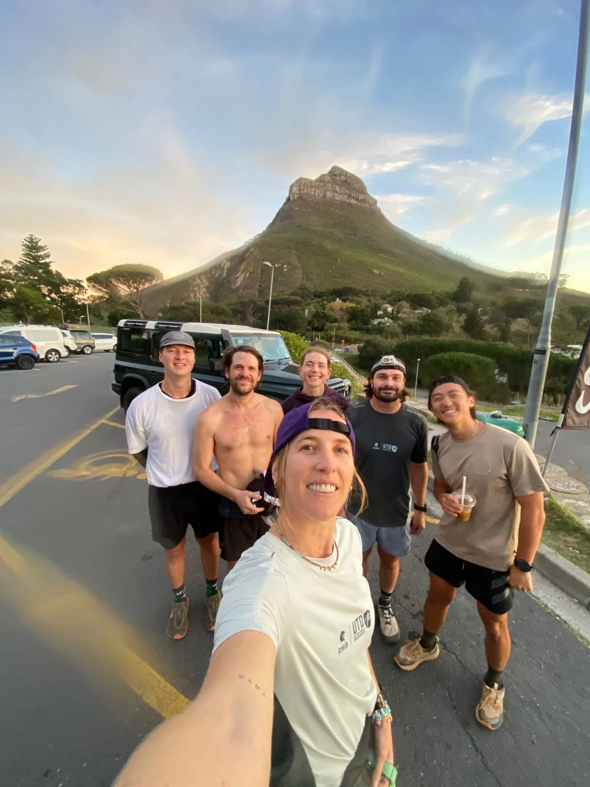 A group of six friends taking a selfie in a parking lot with a tall mountain in the background during sunset.