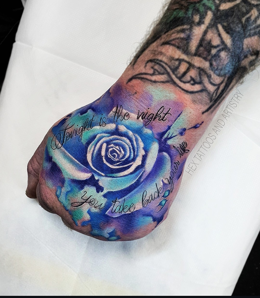 A colorful tattoo of a blue and white rose with the quote, 'Tonight is the night you take back your power,' on a person's forearm.