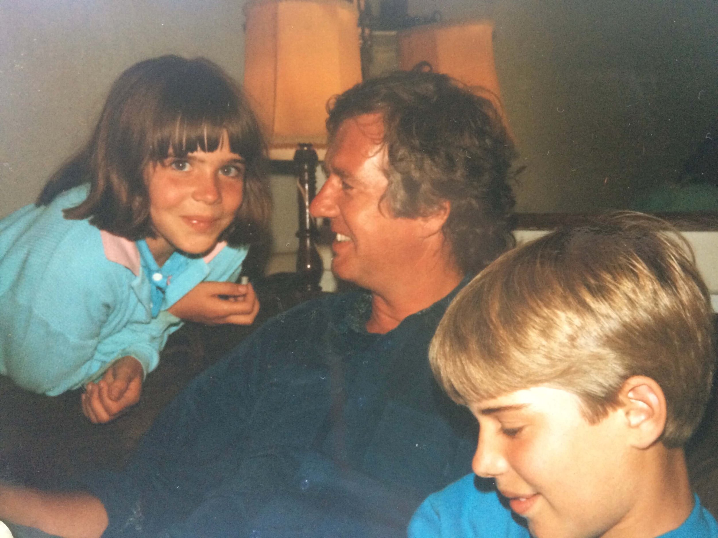 A young girl with brown hair and bangs smiling, leaning close to an adult male with curly hair, both engaged in conversation, indoors with a lamp in the background.