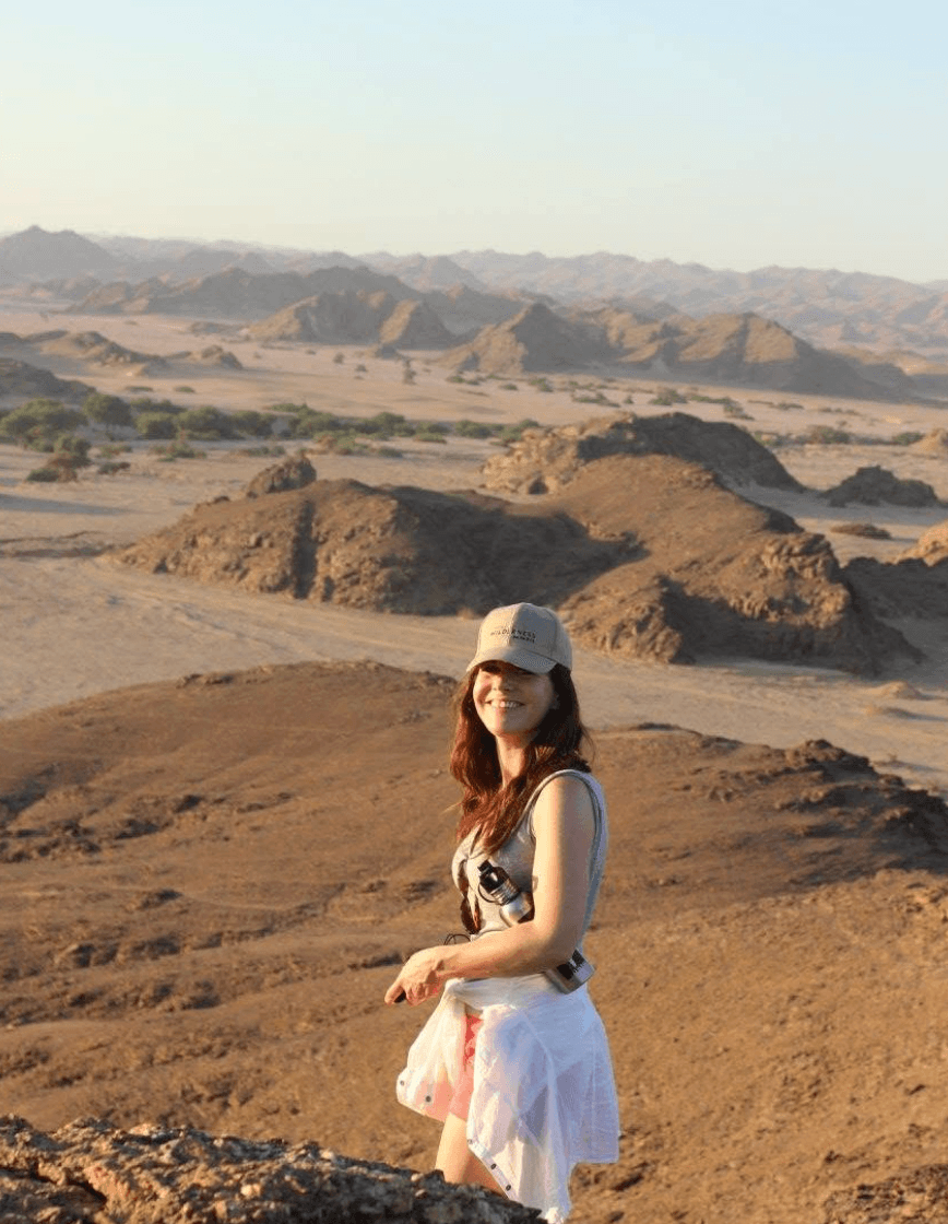 Sally Kirby stands smiling in a desert landscape with rocky formations and distant mountains.