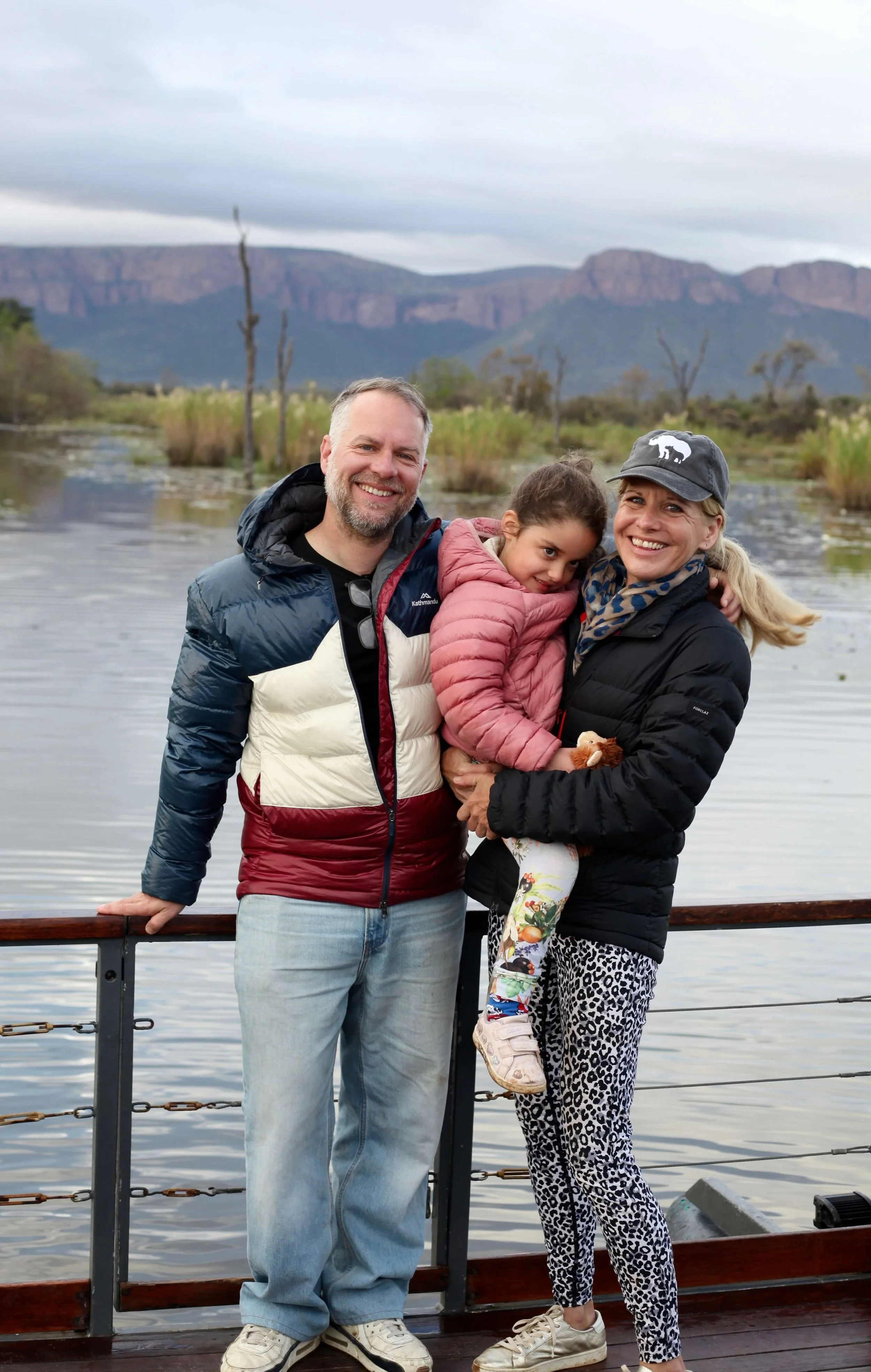 A family of three, a man, a woman, and a young girl, standing on a boat with a lake, trees, and mountains in the background, smiling at the camera.