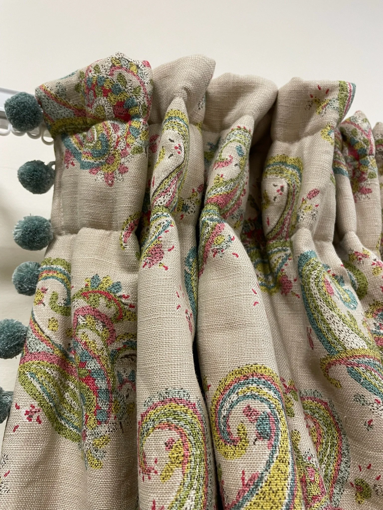 Close-up of a floral patterned curtain with multicolored embroidery and teal pom-pom trim.