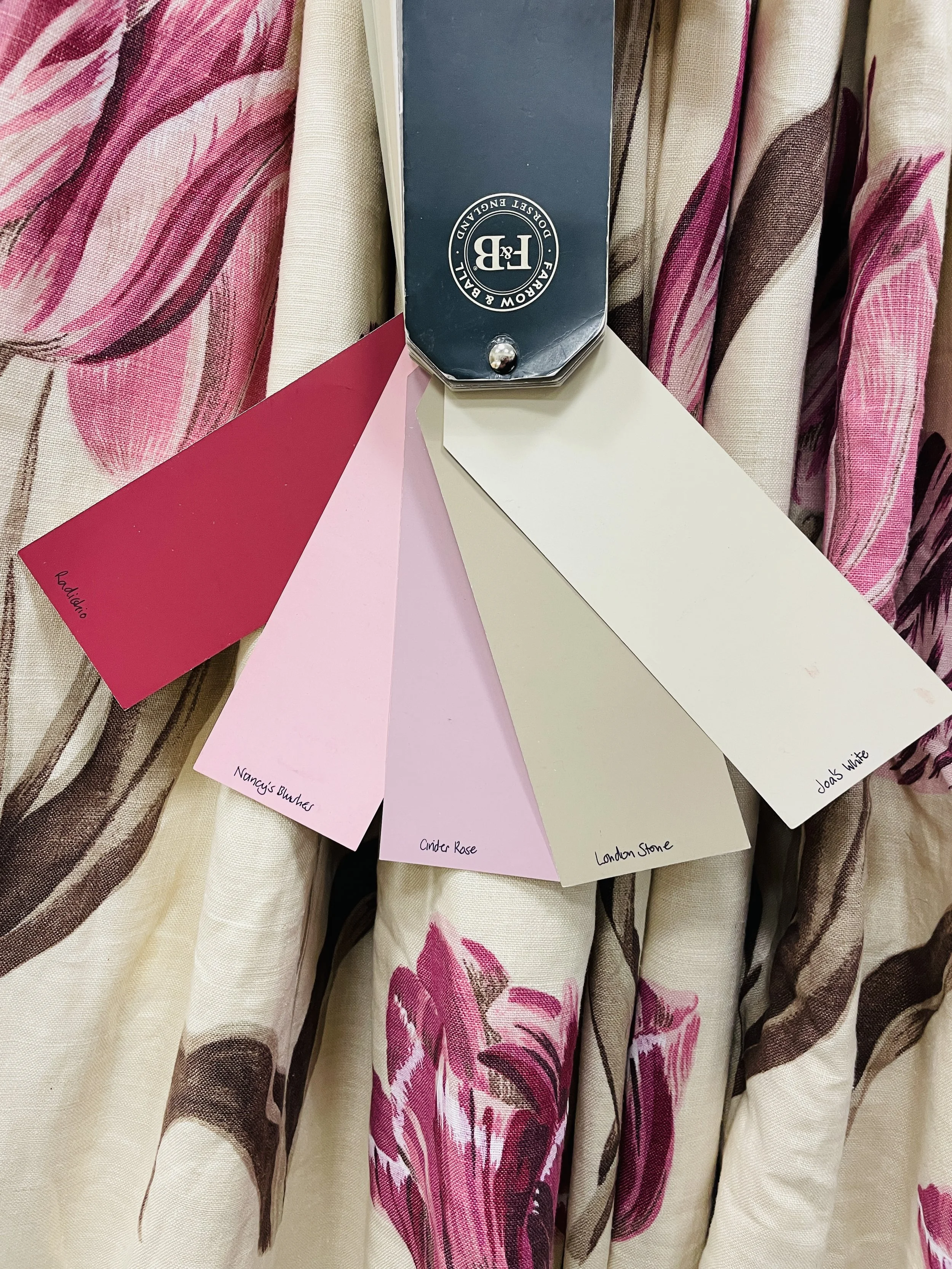 Color swatches of paint samples labeled with names: Ladino, Nancy's Blushes, Cinder Rose, London Stone, Joan White, arranged on floral fabric.