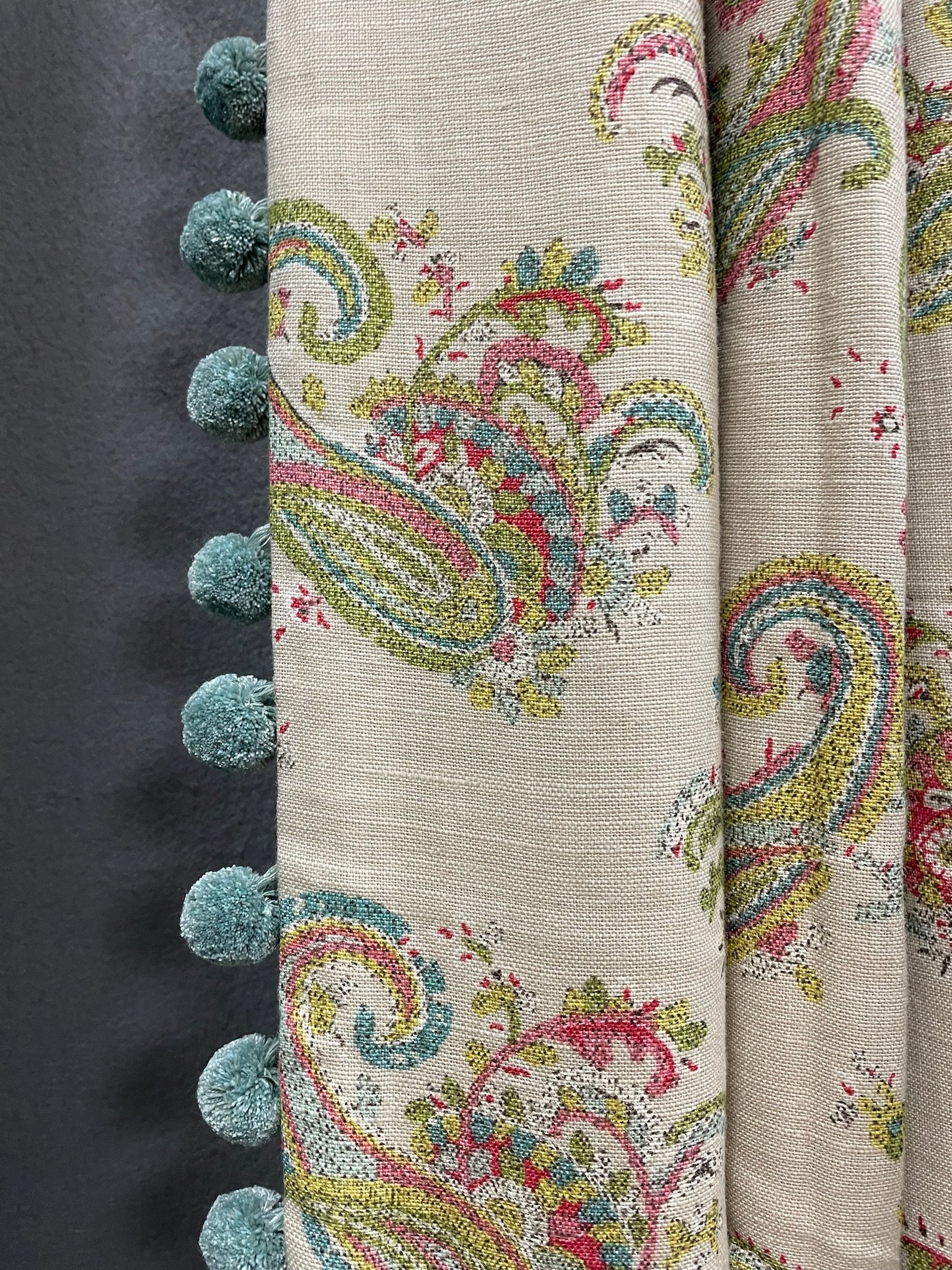 Close-up of embroidered fabric with colorful paisley and floral patterns, adorned with turquoise pom-pom trim along the edge.