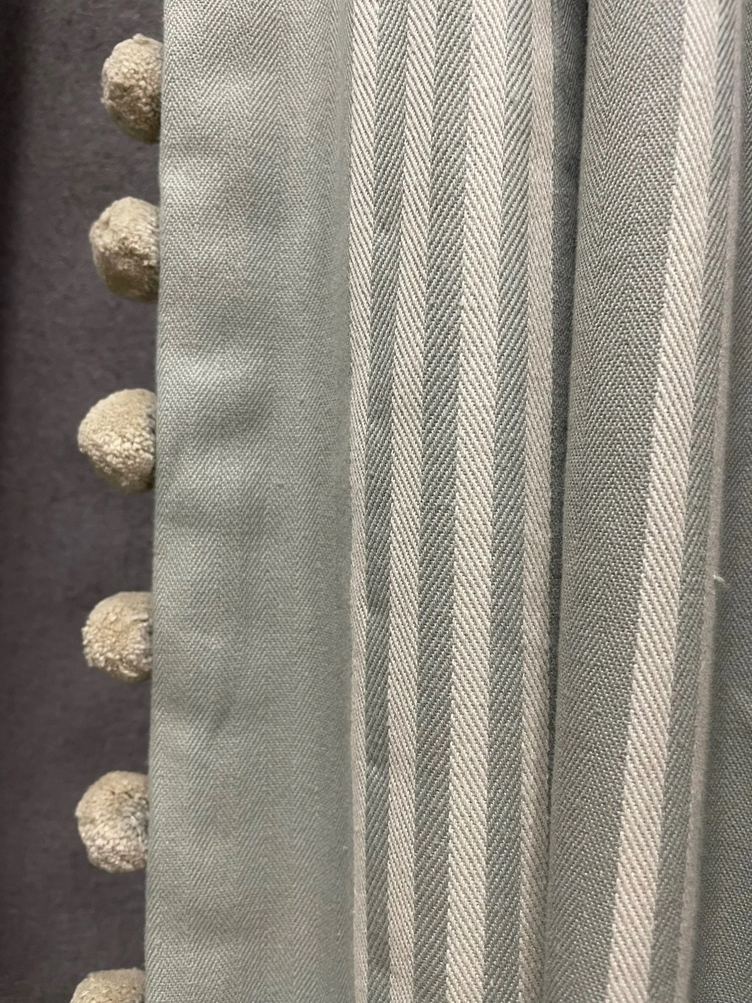Close-up of Blue grey curtain fabric with a herringbone pattern and round decorative pom poms along the edge.