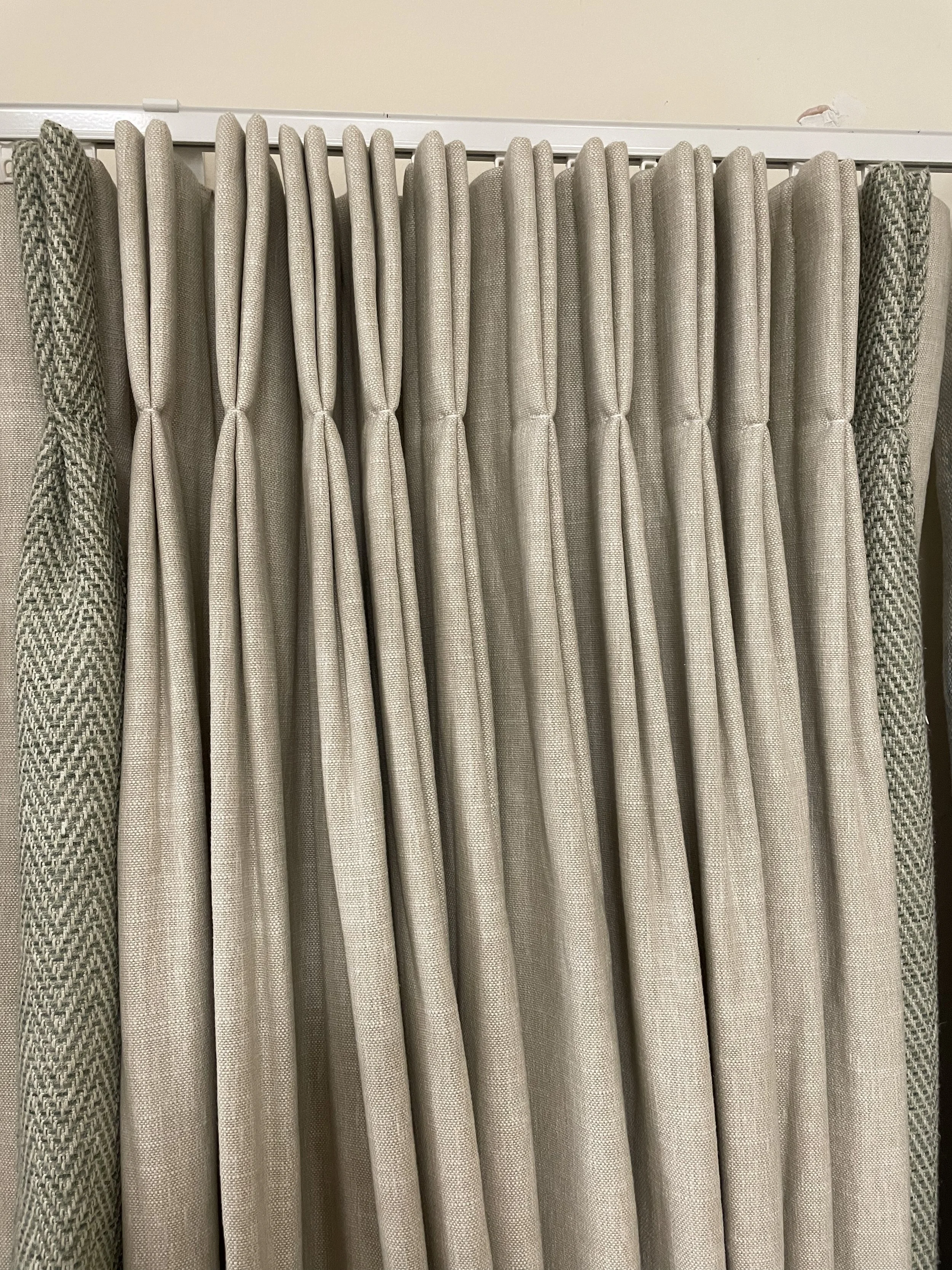 A close-up of beige curtains with a textured, woven pattern, hanging on a white curtain rod against a cream-colored wall.