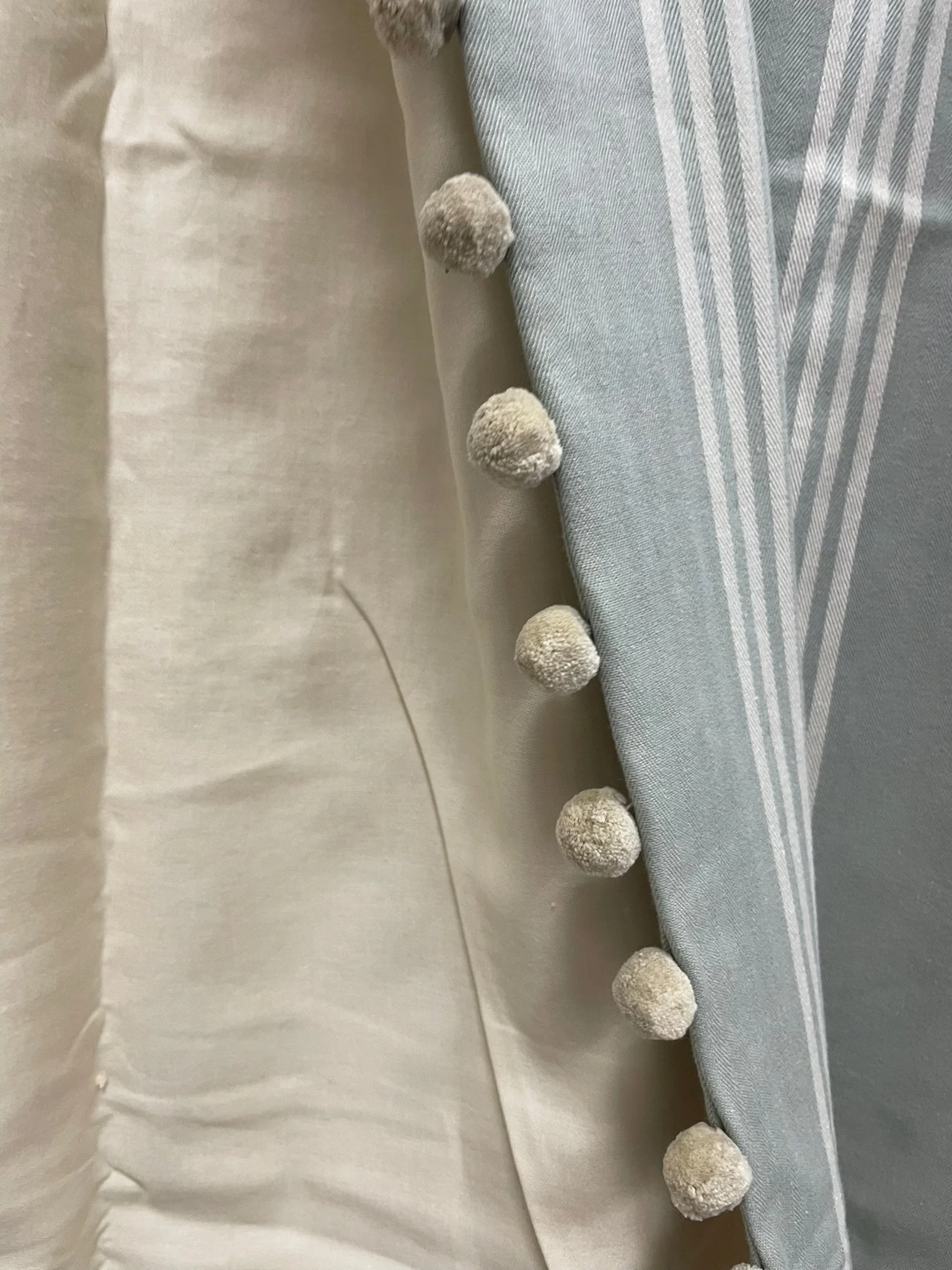 Close-up of a bedsheet with pom-pom trim along the edge, featuring beige pom-poms on a light blue and white striped fabric.