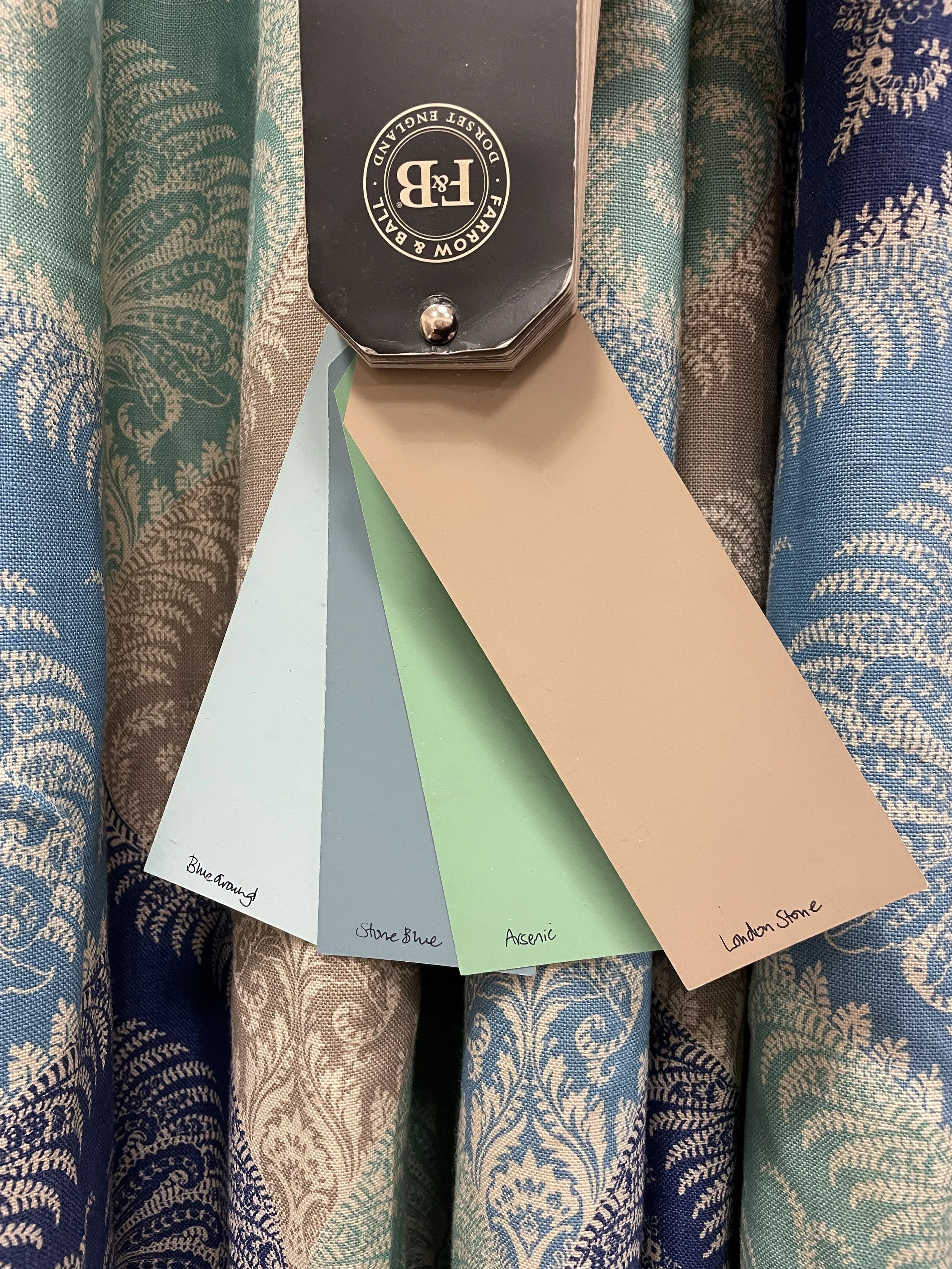 Color swatches labeled Blue Grey, Stone Blue, Arsenic, London Stone, placed on patterned fabric with mixed blue, beige, and green colors.