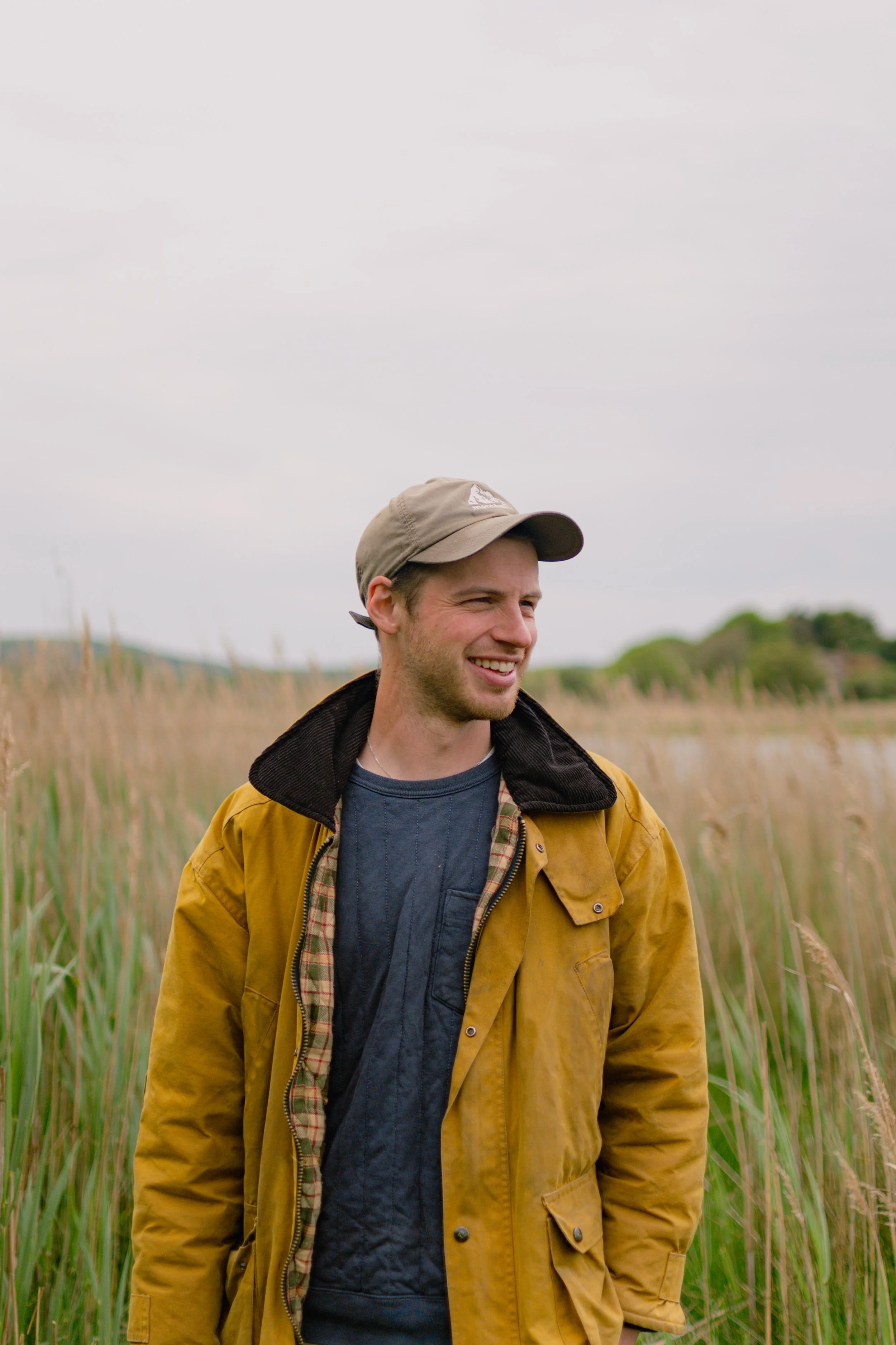 A young man outdoors in a field of tall grass, wearing a beige cap, yellow jacket, and dark shirt, smiling.