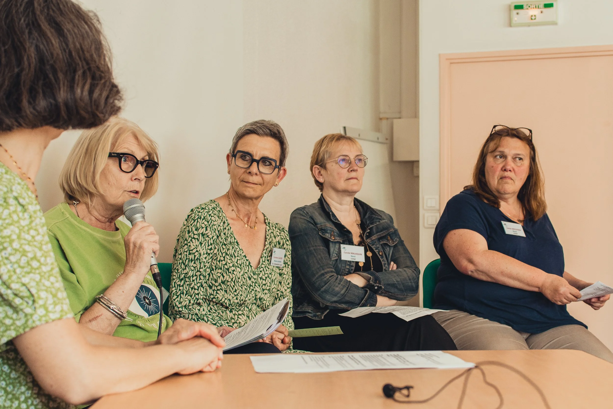 Four women are sitting at a table during a discussion or panel. One woman is speaking into a microphone, while the others listen. They are all wearing name tags and casual clothing.