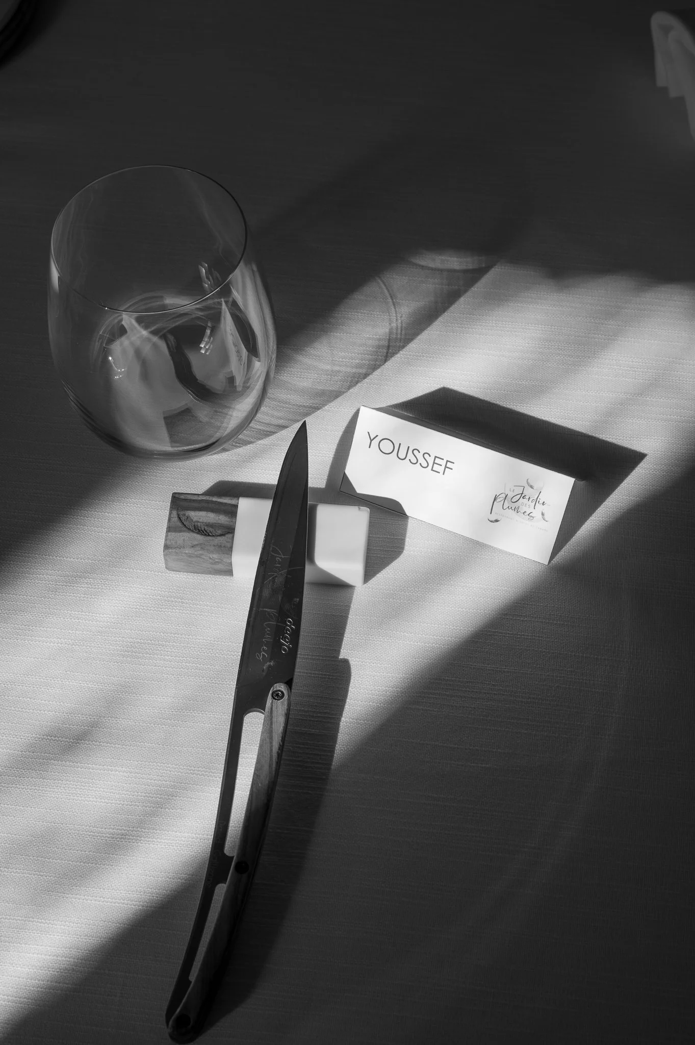 A table setting with a wine glass, a chef's knife, a name card reading 'Youssef,' a small ceramic dish, and a folded napkin, with sunlight casting shadows.