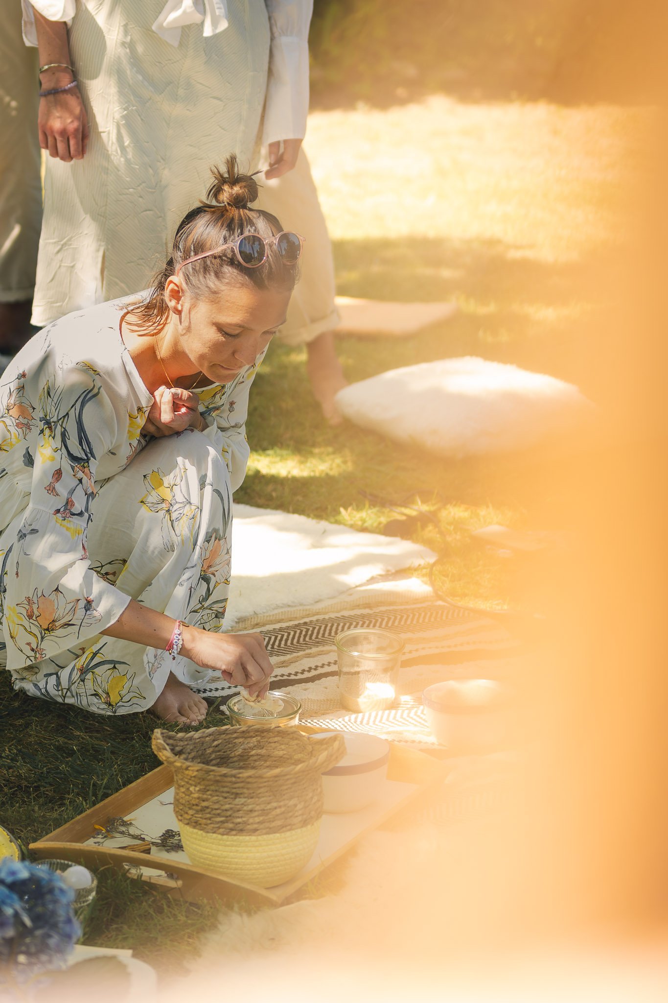 Une femme en tenue légère s'agenouille en prière ou méditation lors d'une cérémonie en plein air, avec d'autres personnes en arrière-plan.