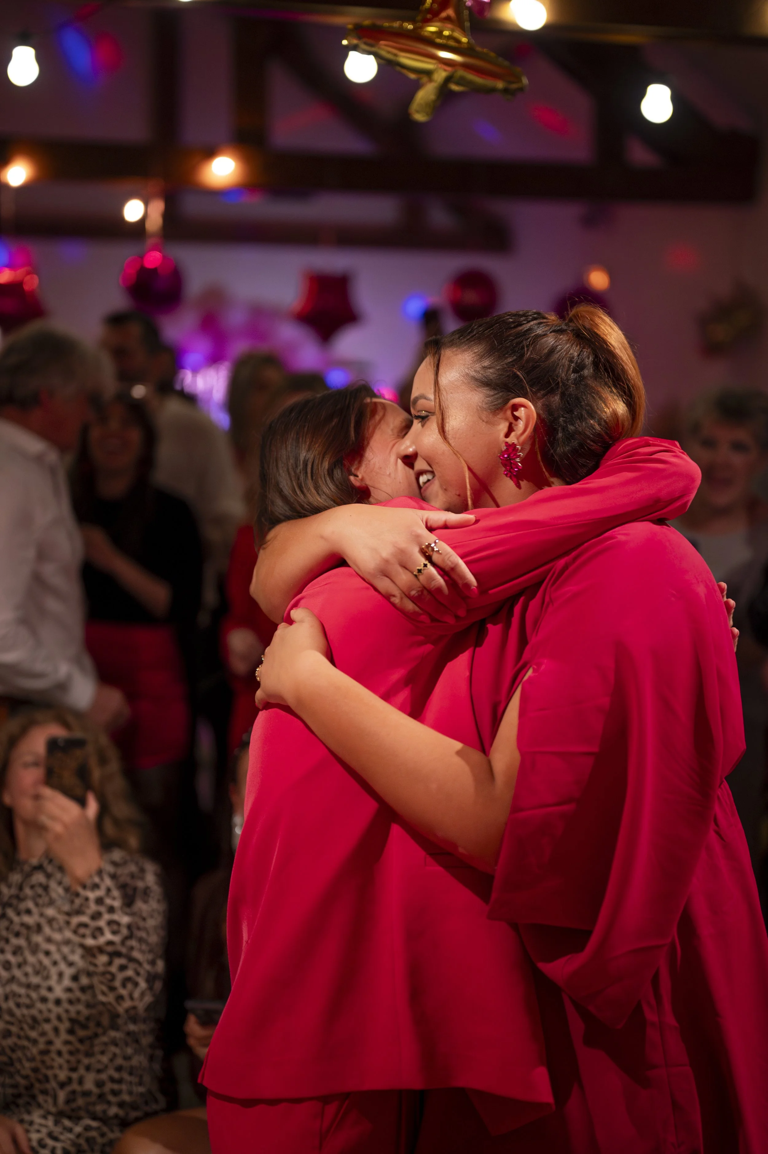 Two women hugging at a celebration or party, surrounded by other people, with festive decorations and pink lighting in the background.