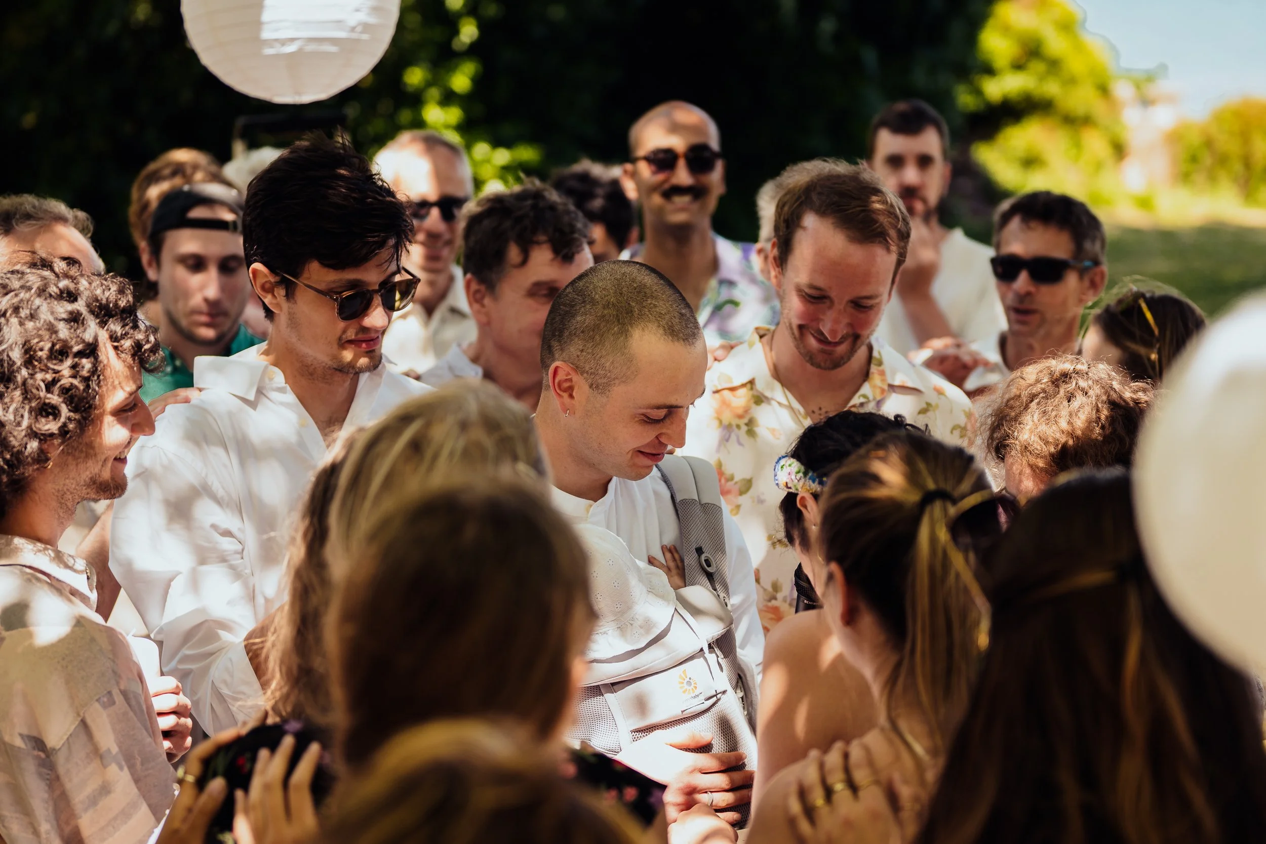 A group of people gathered outdoors, with the focus on a young man with a shaved head who appears to be the center of attention, surrounded by smiling friends and family, during daylight.