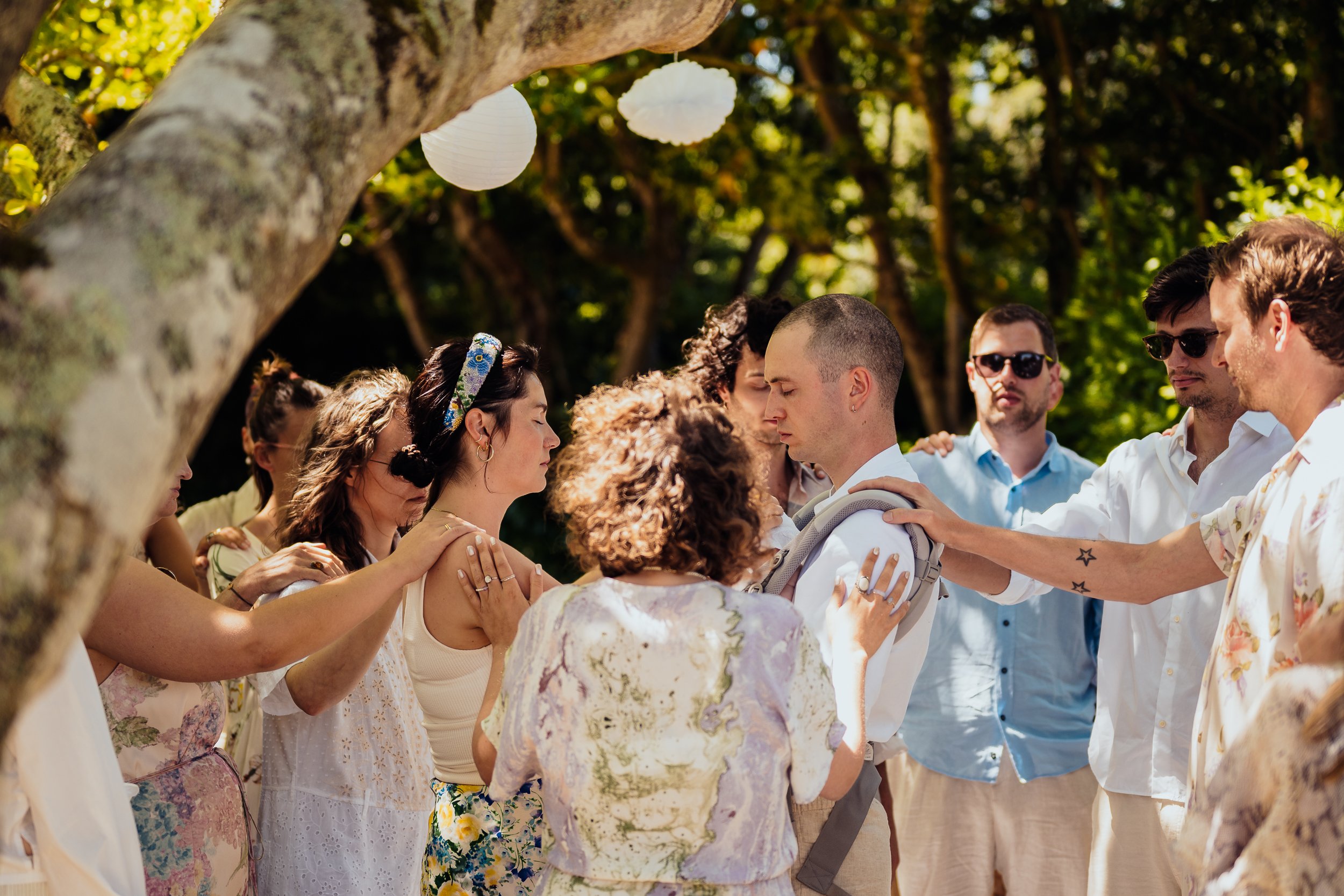 Group of people praying or performing a ritual outdoors under a tree, with white hanging lanterns in the background.