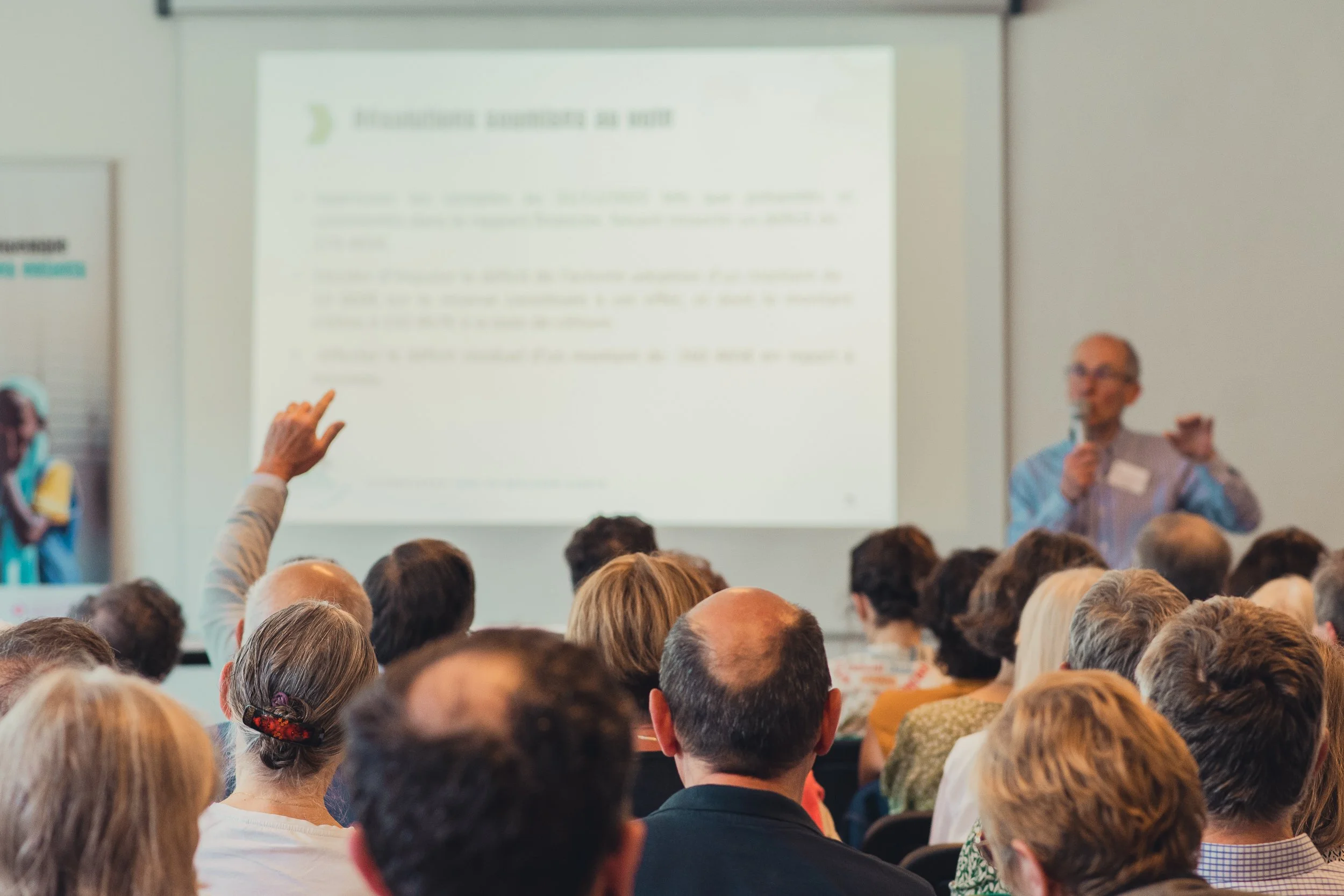A speaker giving a presentation to an audience in a conference room with a large screen behind him. Audience members are listening attentively, with one person raising their hand.