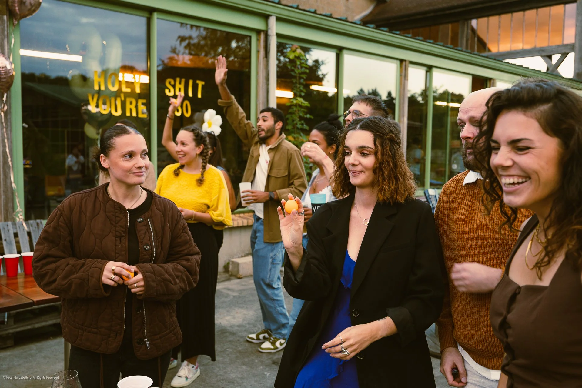 Groupe de personnes souriantes lors d'une fête en extérieur, certains jouent à des jeux de bière avec des balles en caoutchouc, une femme en avant tient une balle orange, en arrière une fenêtre avec la phrase humoristique "HOLY SHIT YOU'RE SO"
