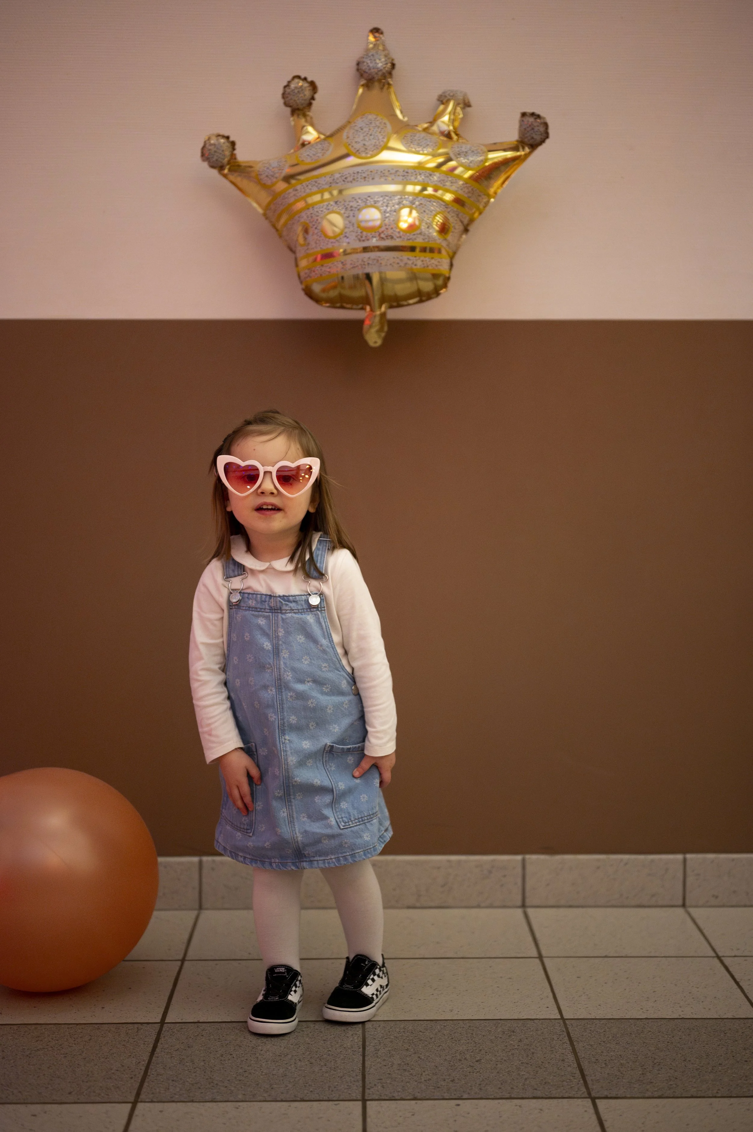 A young girl wearing heart-shaped pink sunglasses and a denim dressoveralls, standing indoors on tile flooring with a large gold balloon in the shape of a crown hovering above her, and a gold exercise ball on the floor nearby.